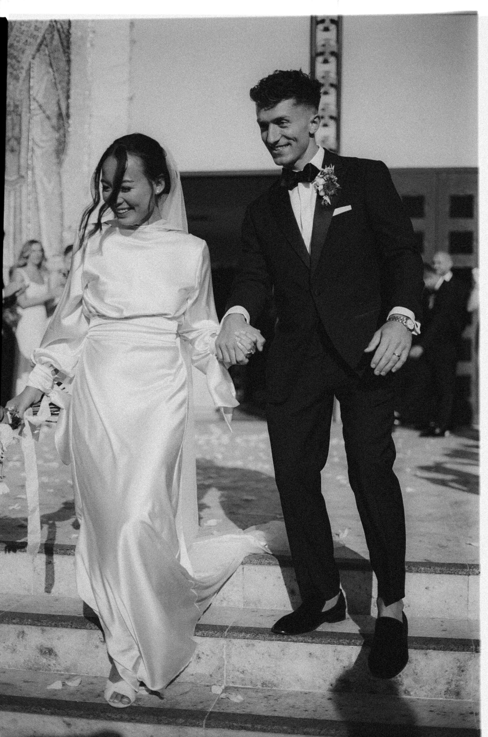 A newlywed couple in formal attire walks down steps, smiling and holding hands. The bride wears a long dress and veil, while the groom is in a suit and bow tie.