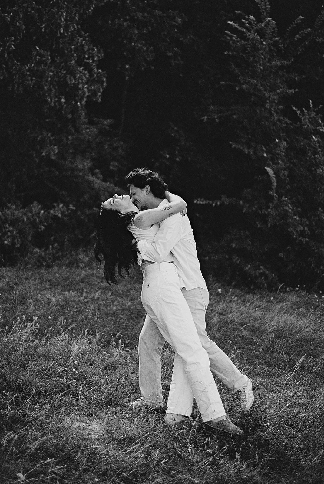 A couple embraces and dances in a grassy field, dressed in light-colored clothing. The image is in black and white during their candid engagement photos