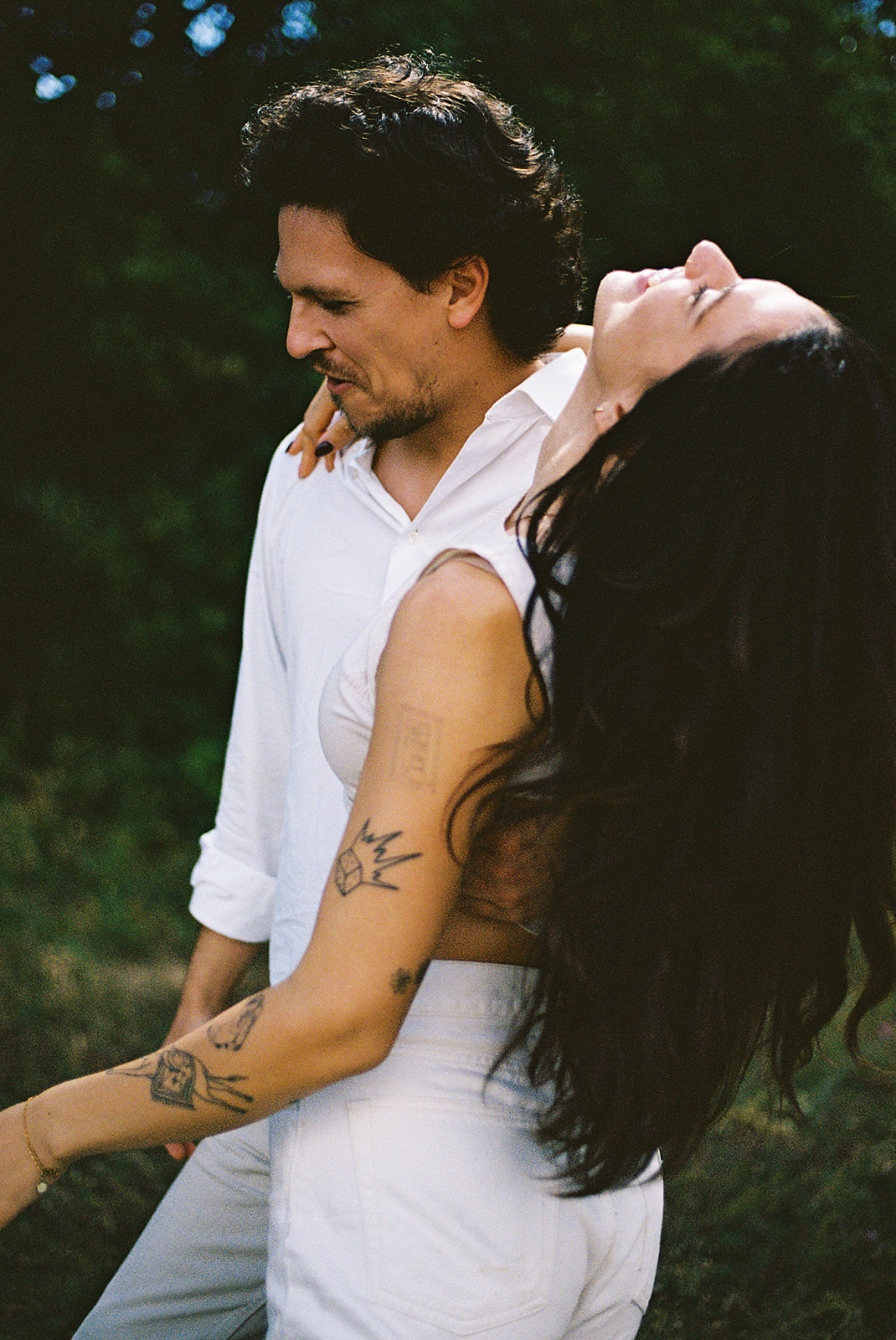 A couple dressed in white embraces outdoors, with greenery in the background during their candid engagement photos