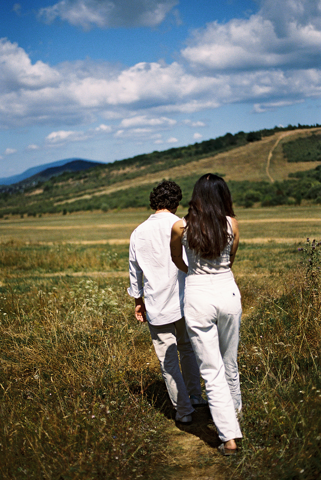 A couple dressed in white embraces outdoors, with greenery in the background during their candid engagement photos