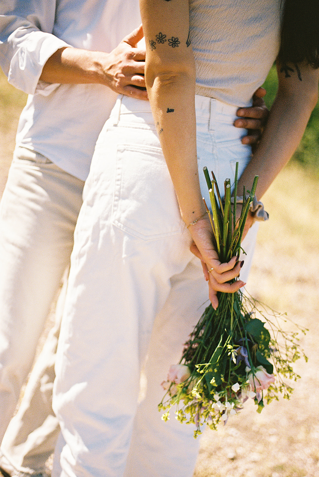 A couple dressed in white embraces outdoors, with greenery in the background during their candid engagement photos