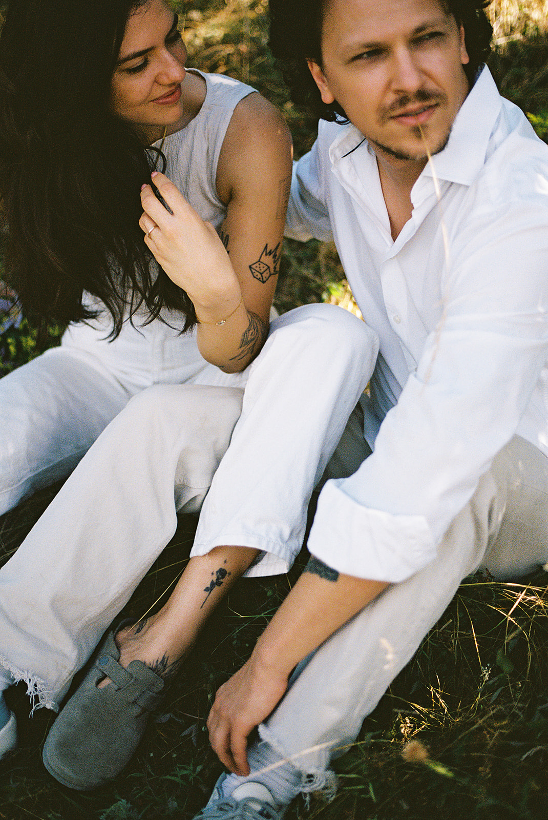 A couple dressed in white embraces outdoors, with greenery in the background during their candid engagement photos