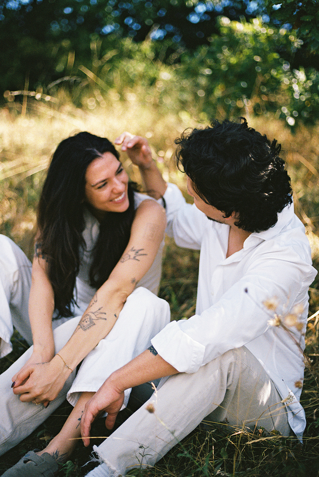 A couple dressed in white embraces outdoors, with greenery in the background during their candid engagement photos