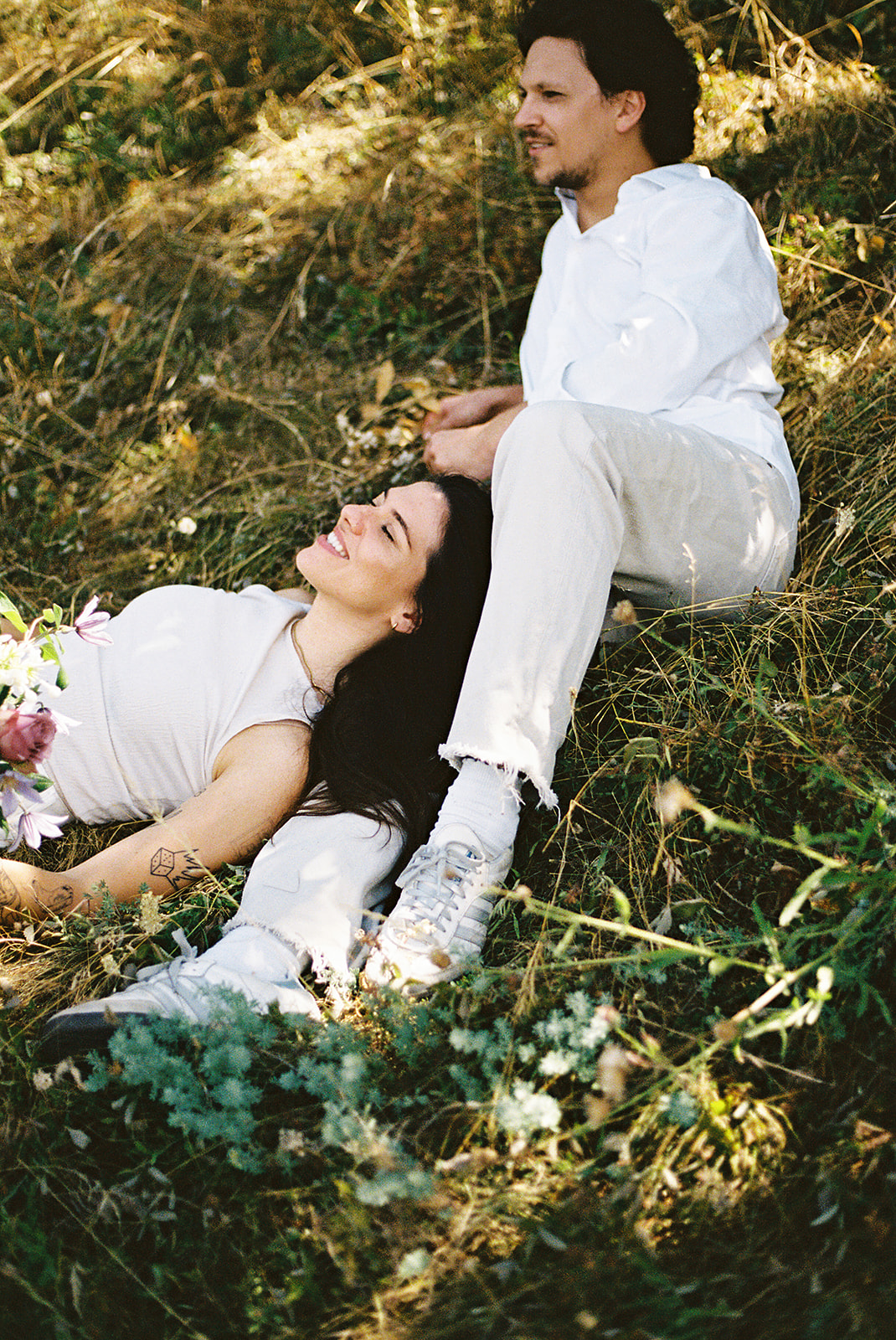 A couple dressed in white embraces outdoors, with greenery in the background during their candid engagement photos