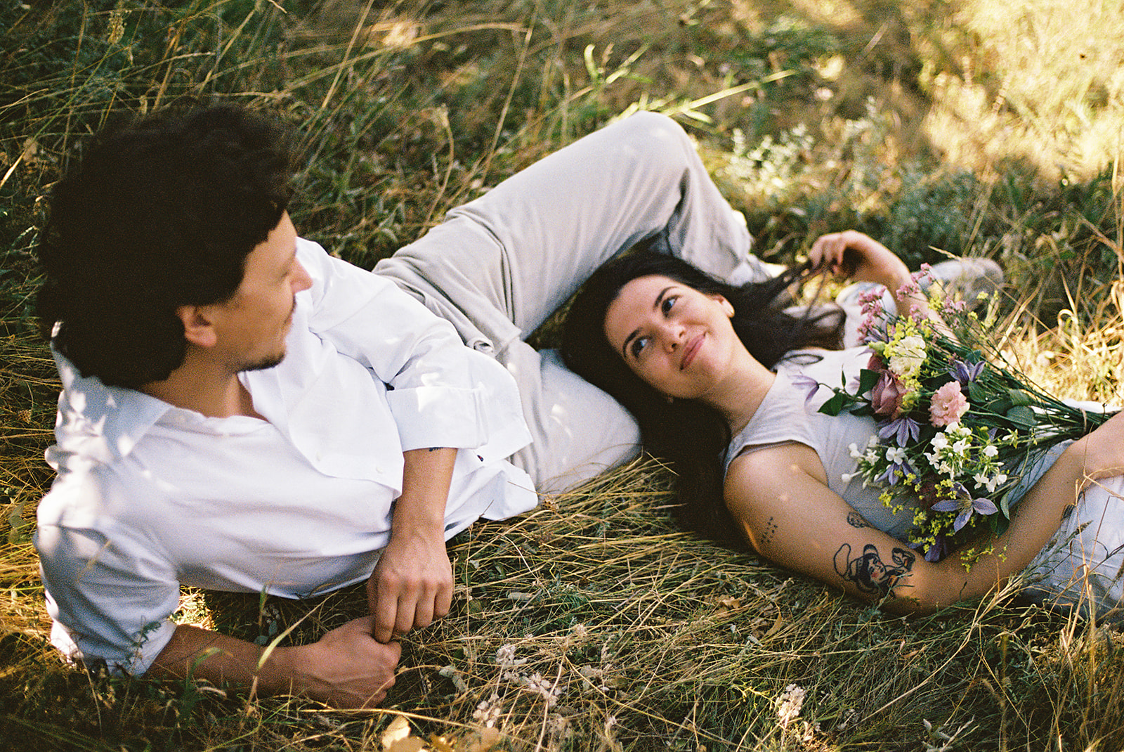 A couple dressed in white embraces outdoors, with greenery in the background during their candid engagement photos