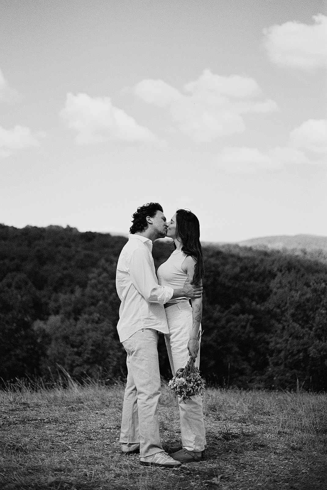 A couple dressed in white embraces outdoors, with greenery in the background during their candid engagement photos