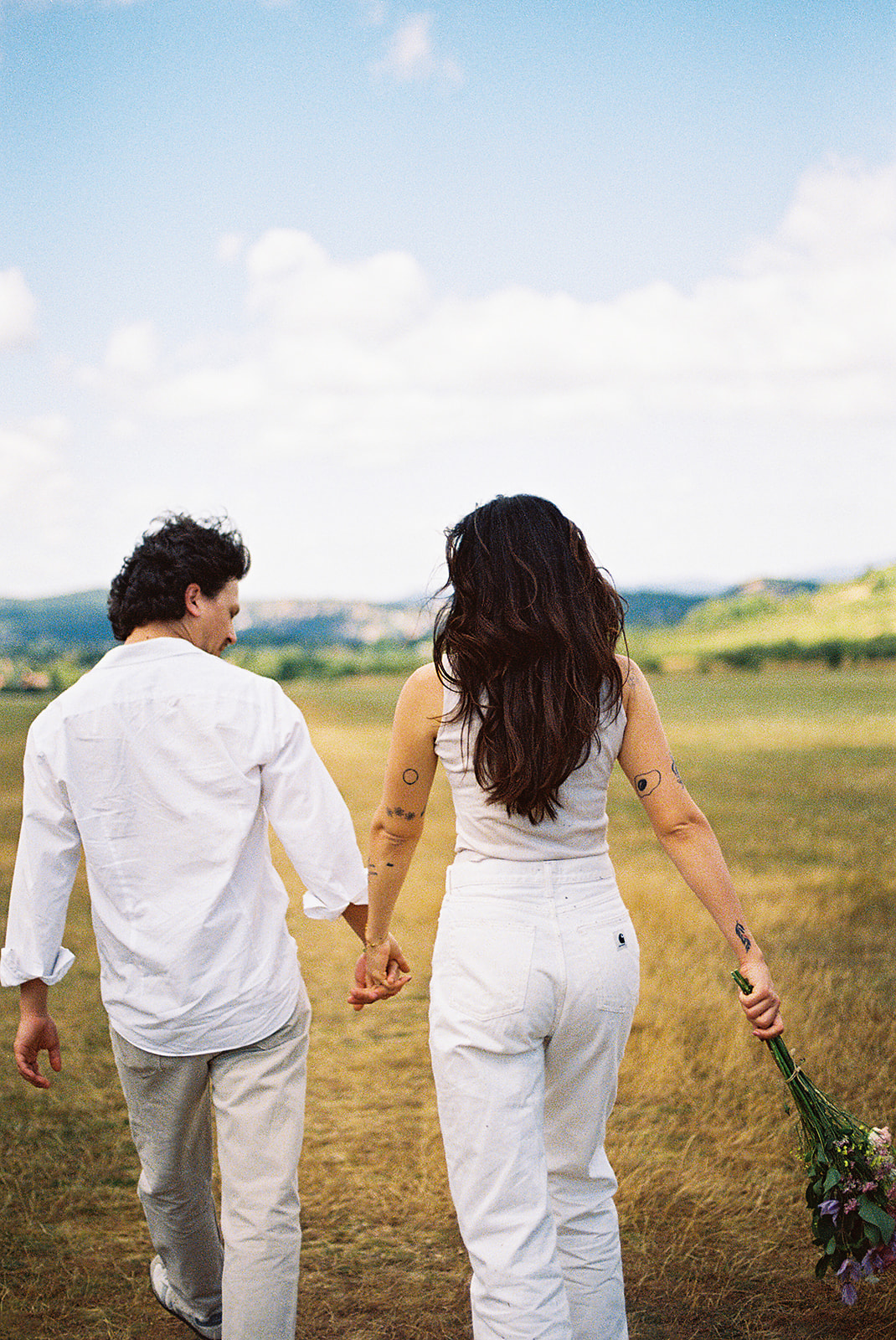 A couple dressed in white embraces outdoors, with greenery in the background during their candid engagement photos