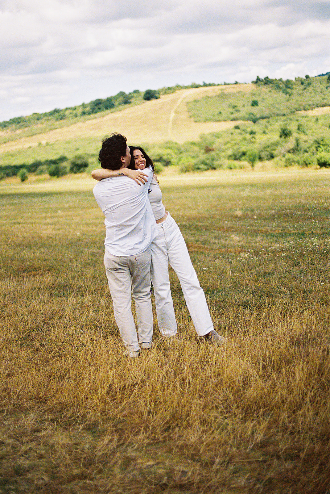 A couple dressed in white embraces outdoors, with greenery in the background during their candid engagement photos