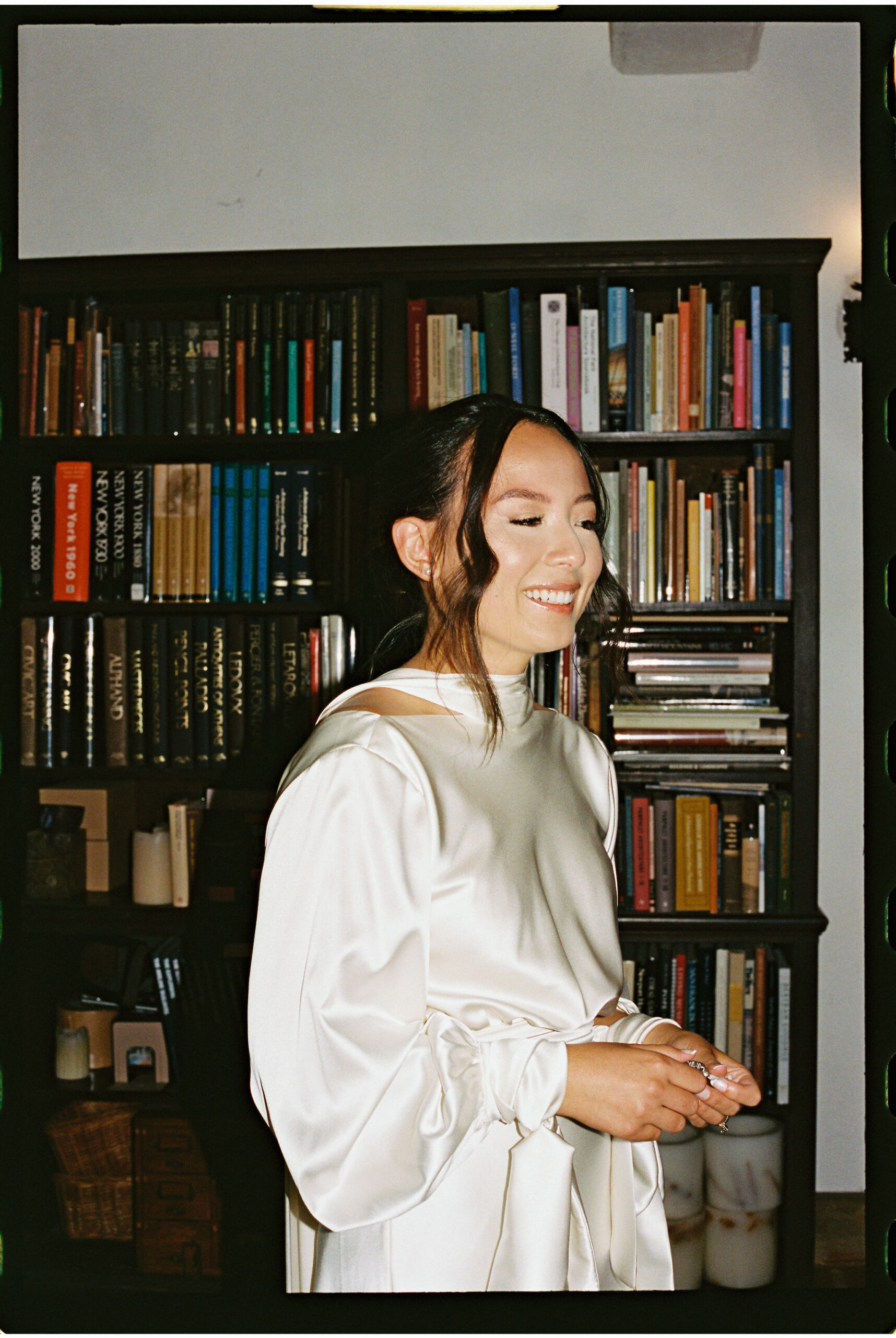 A woman wearing a satin dress stands in front of a bookshelf, smiling with eyes closed.