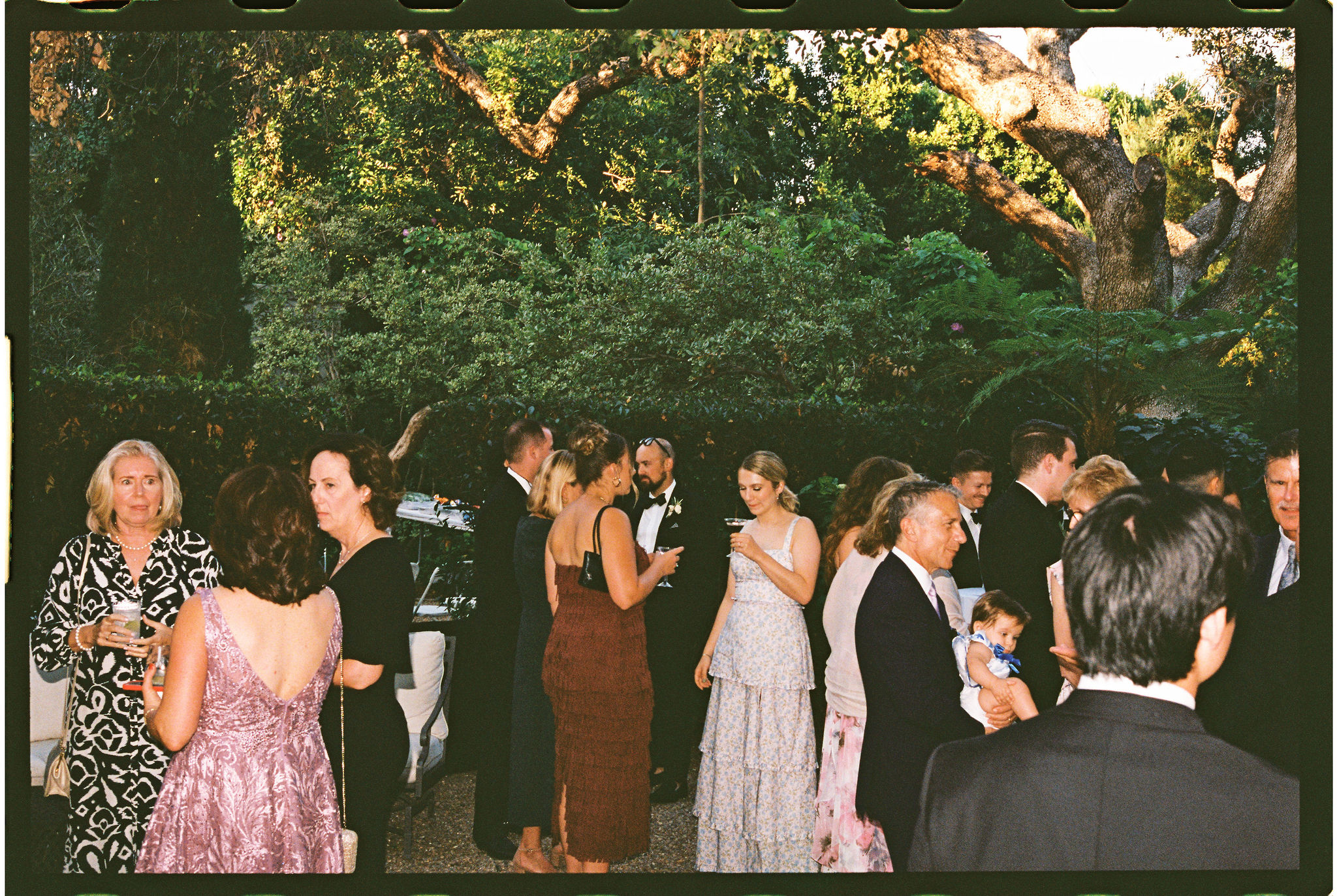 People gathered outdoors at an elegant event, wearing formal attire. Trees and greenery in the background.