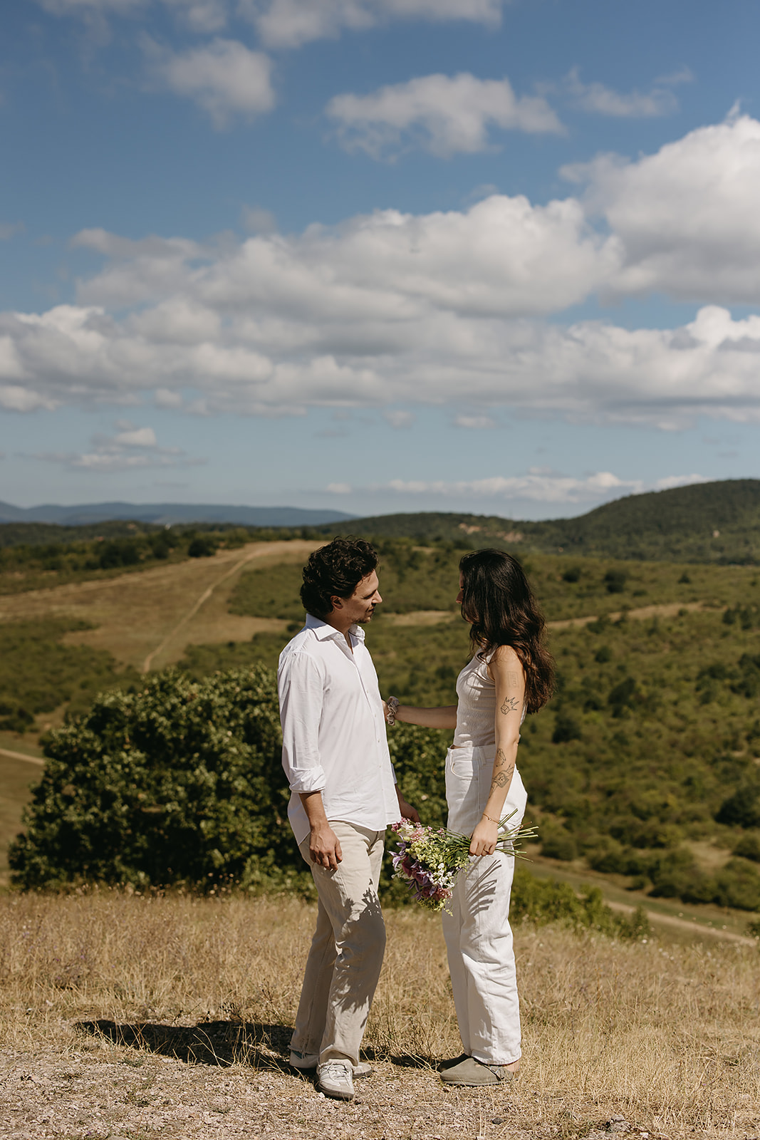 A couple dressed in white embraces outdoors, with greenery in the background during their candid engagement photos