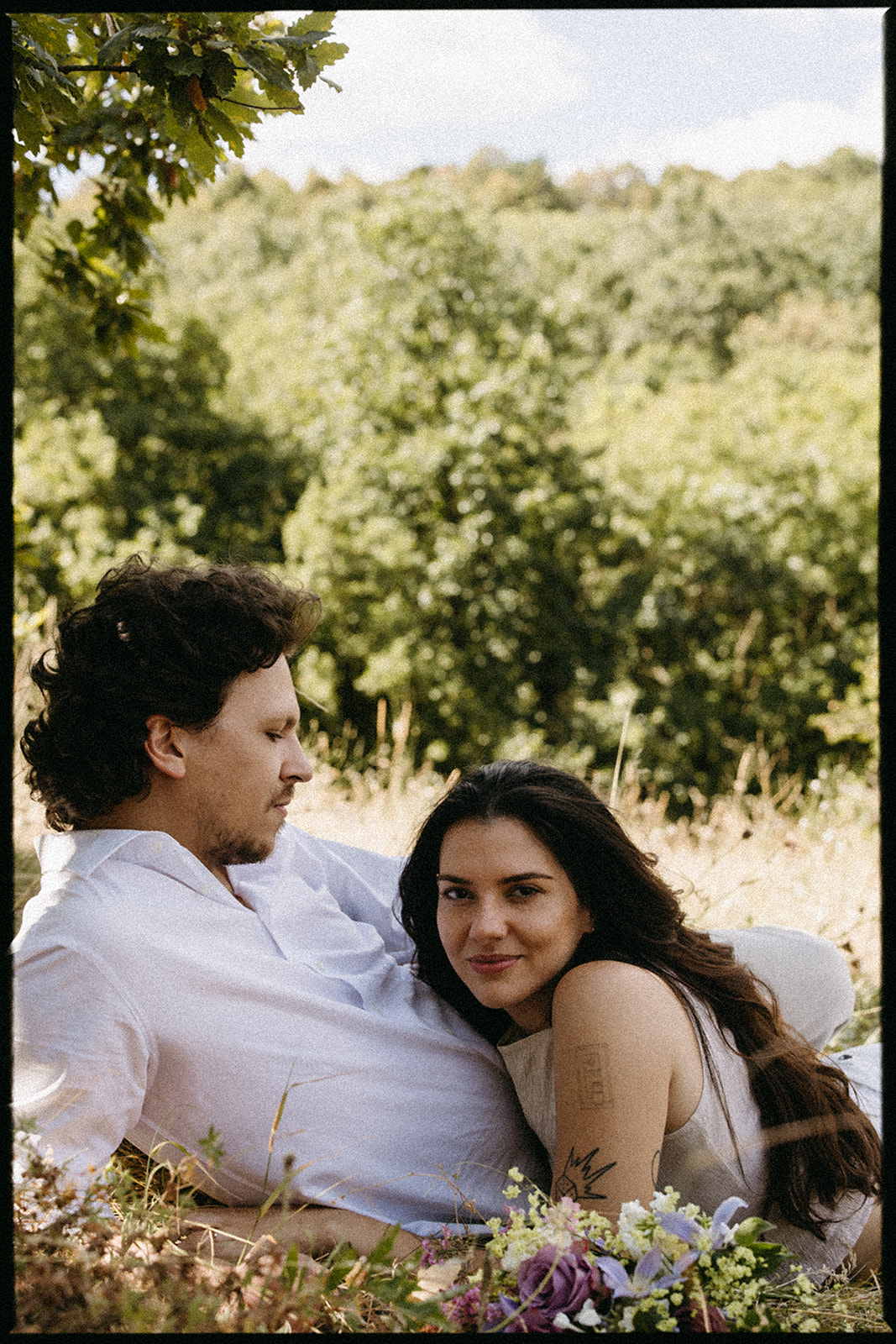 A couple dressed in white embraces outdoors, with greenery in the background during their candid engagement photos