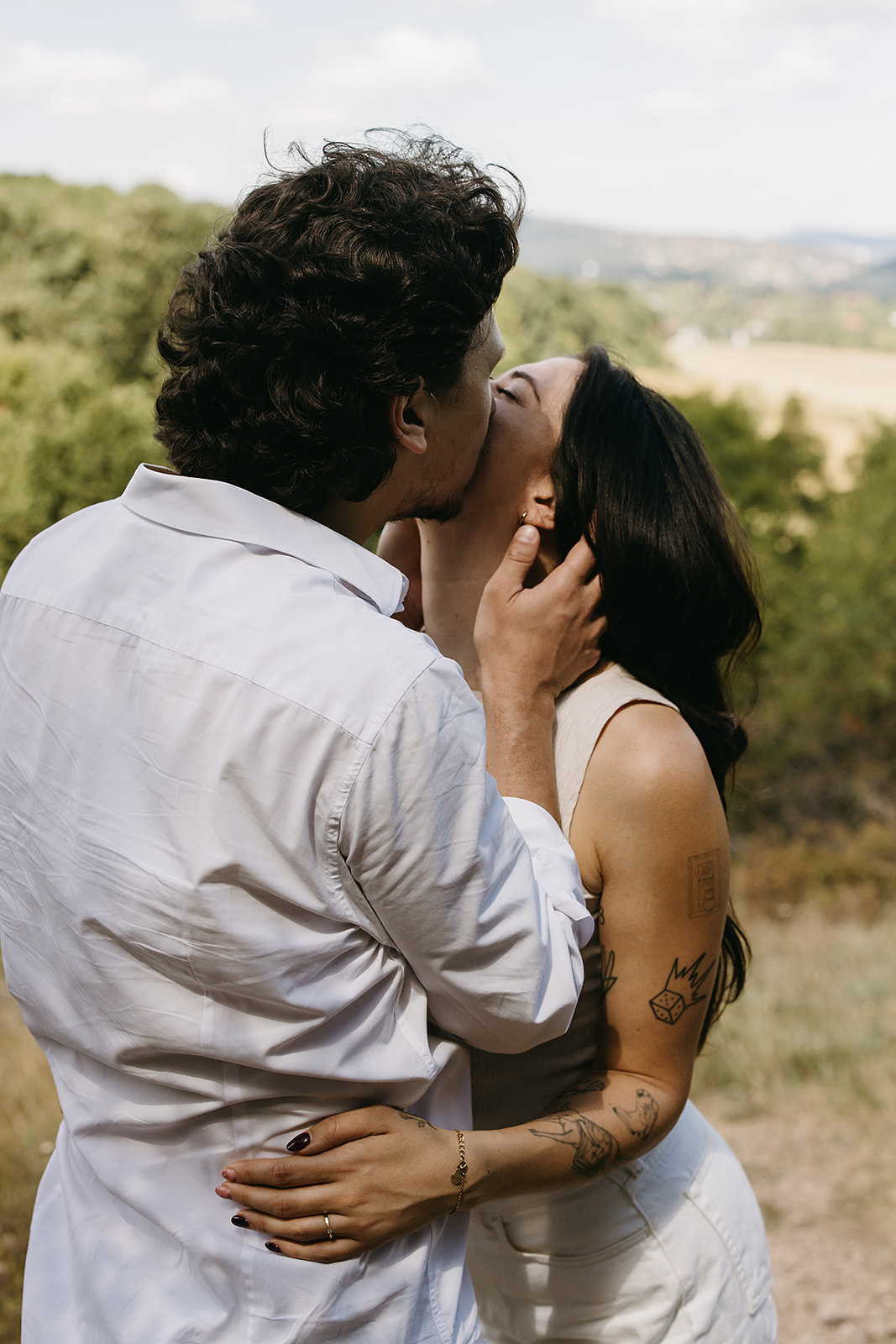 A couple dressed in white embraces outdoors, with greenery in the background during their candid engagement photos