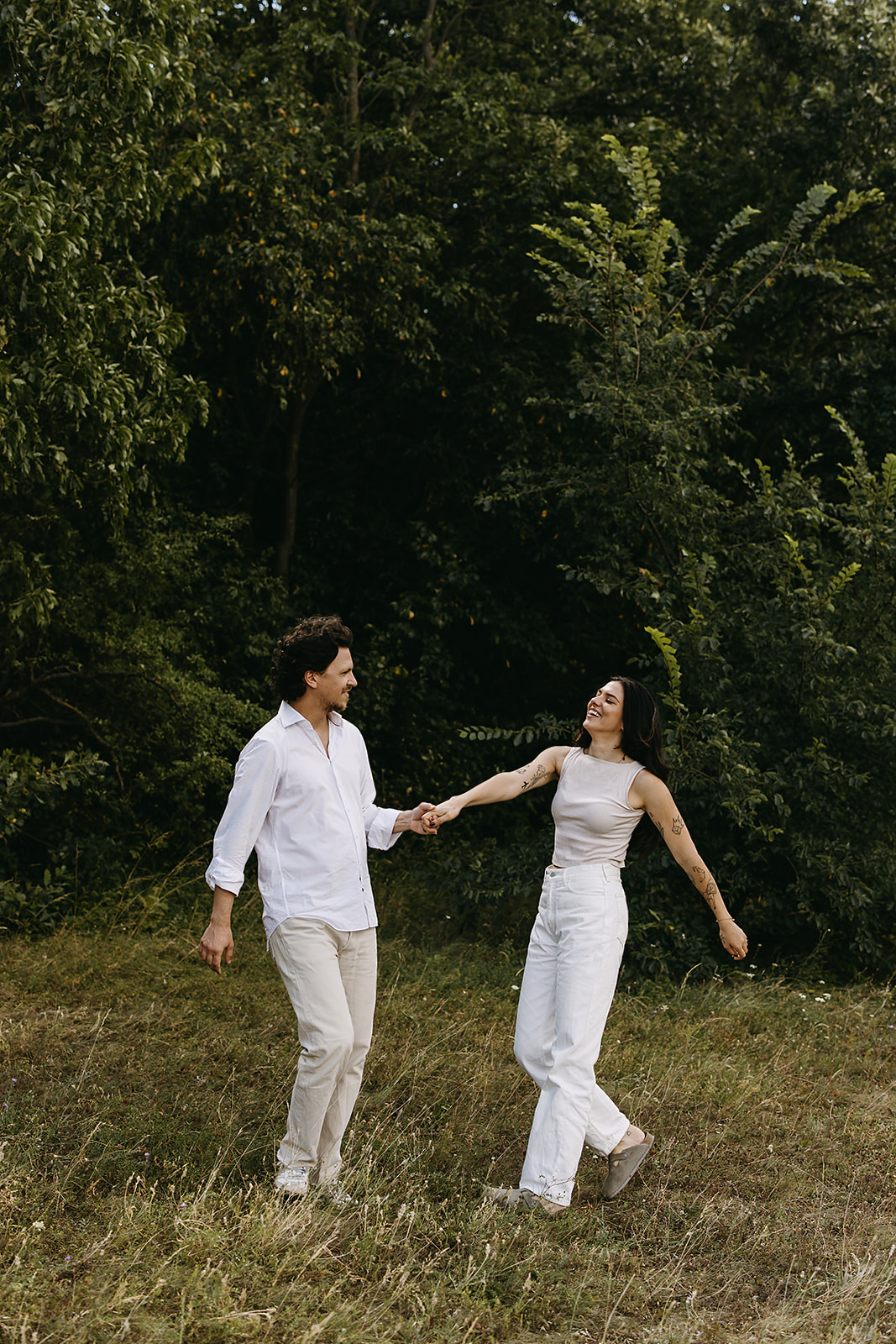 A man and woman holding hands and walking on grass in front of a wooded background.