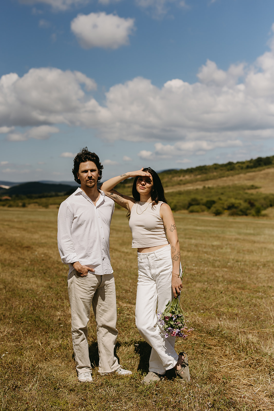 A man and woman stand in an open field under a partly cloudy sky. The woman shields her eyes with her hand and holds flowers. Both wear casual white clothing.