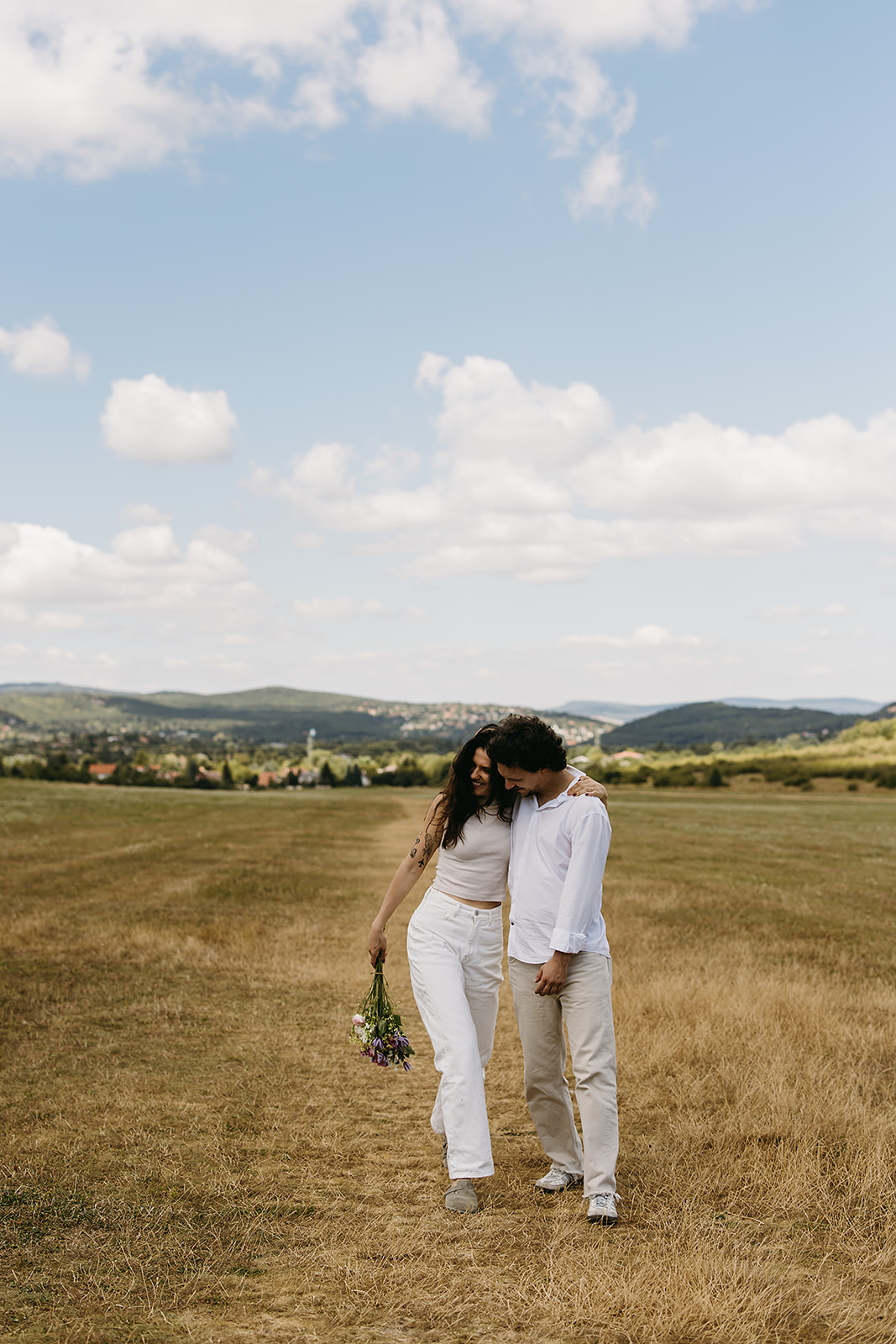 A man and woman stand in an open field under a partly cloudy sky. The woman shields her eyes with her hand and holds flowers. Both wear casual white clothing.