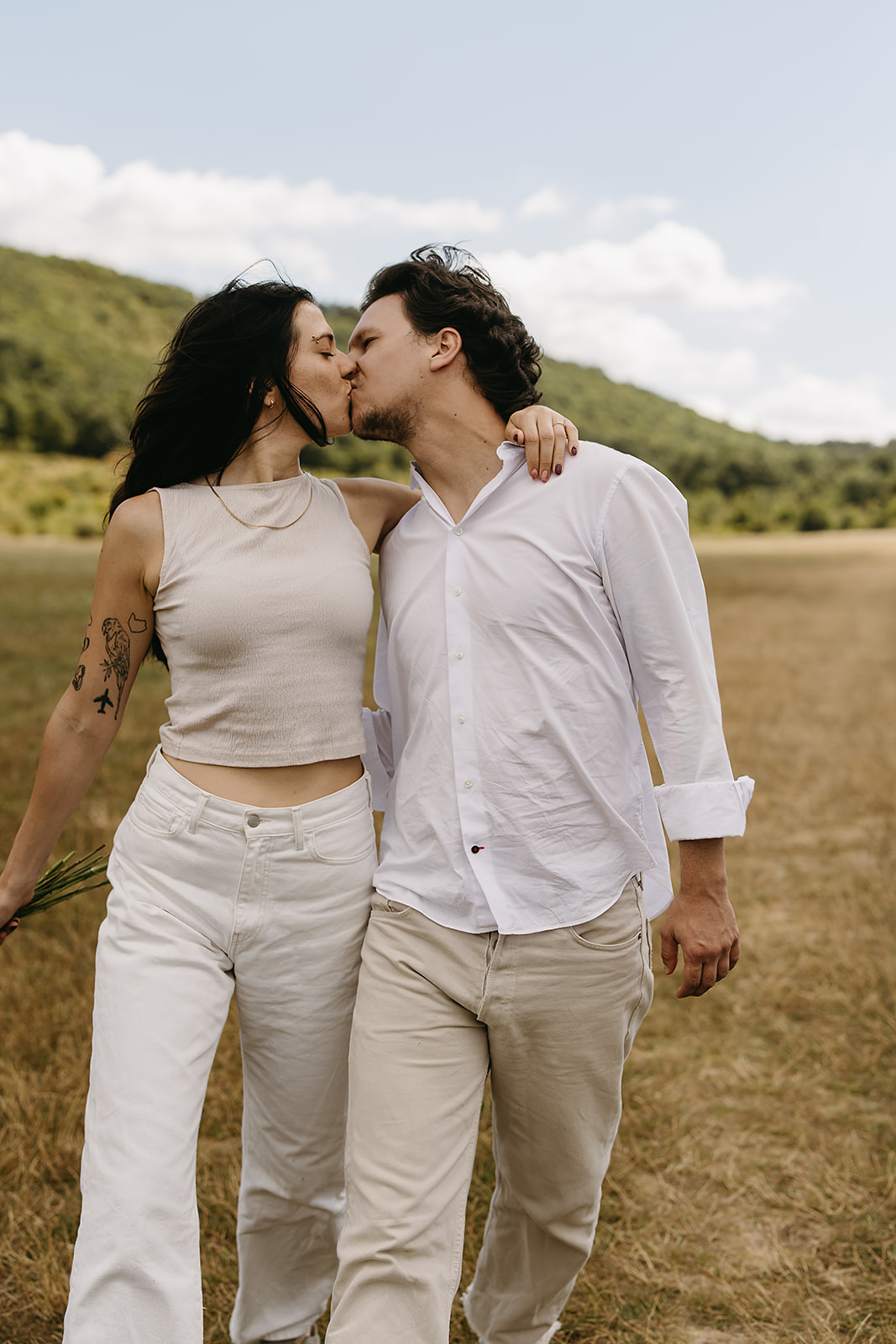 A man and woman stand in an open field under a partly cloudy sky. The woman shields her eyes with her hand and holds flowers. Both wear casual white clothing.