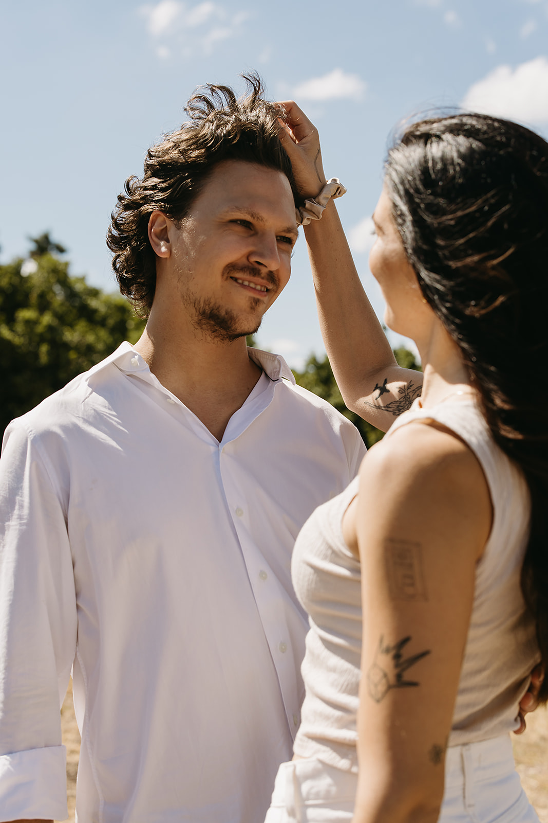 A couple dressed in white embraces outdoors, with greenery in the background during their candid engagement photos