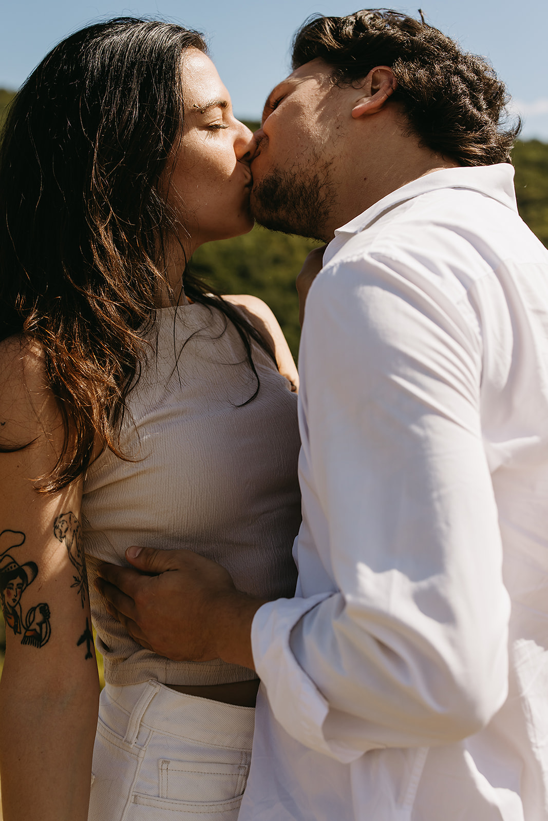 A couple dressed in white embraces outdoors, with greenery in the background during their candid engagement photos