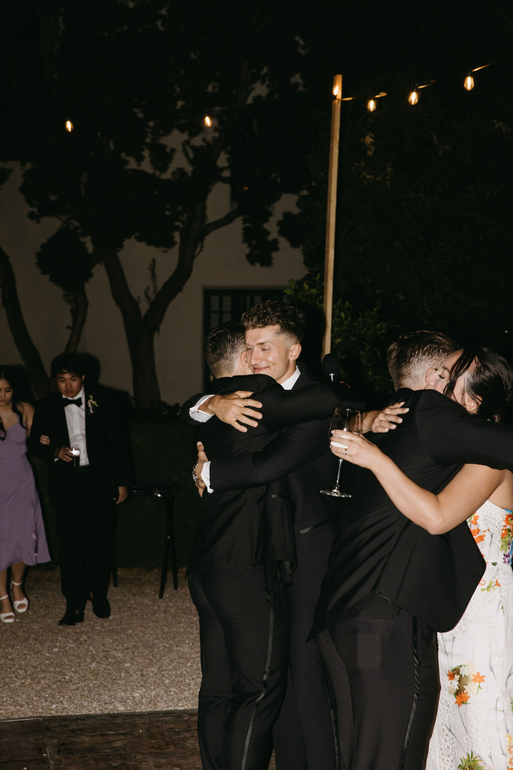 Four people in formal attire are hugging at an outdoor event, with two others in the background. One holds a wine glass. String lights are visible above.