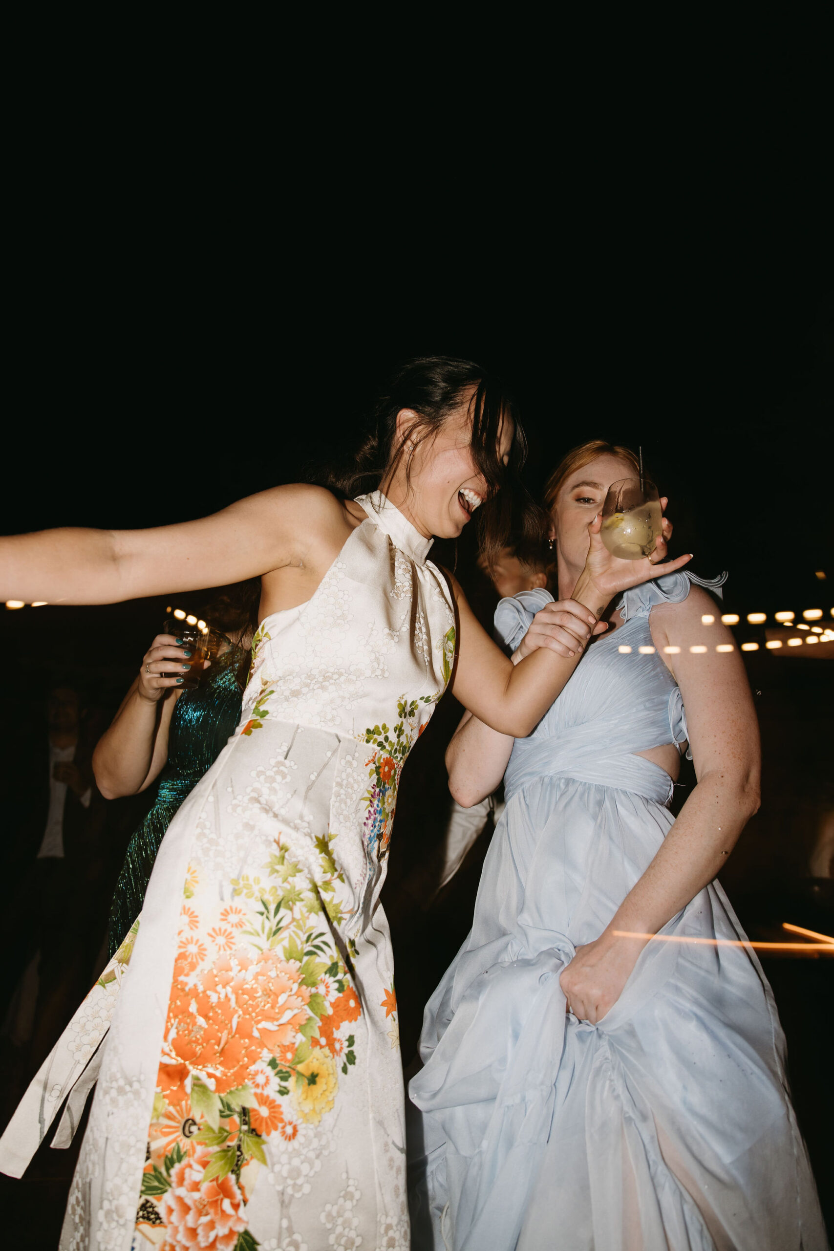 Two women in formal dresses are joyfully dancing and holding drinks at a nighttime event.