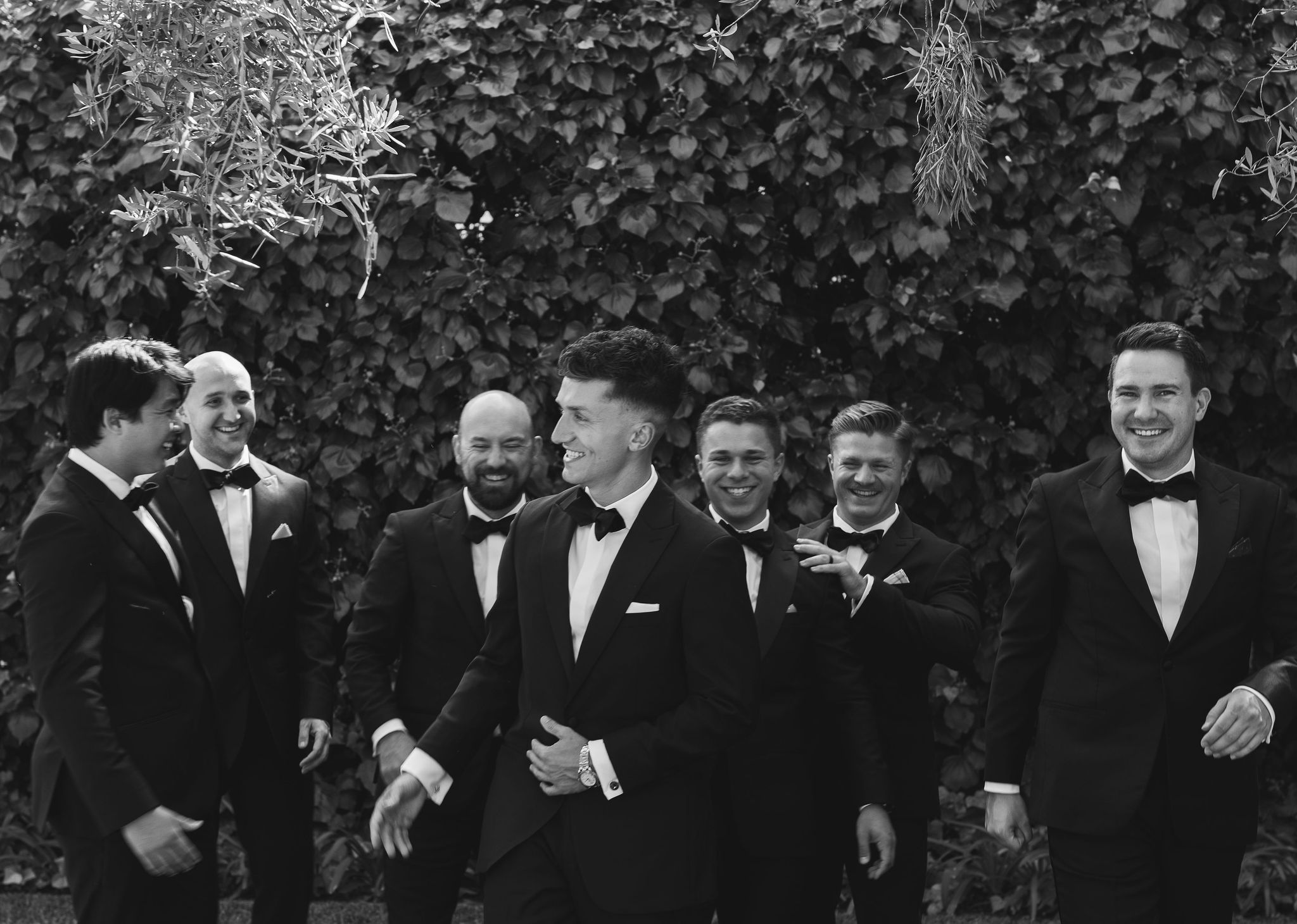 A group of men in tuxedos laugh and converse outdoors, with ivy-covered walls in the background.