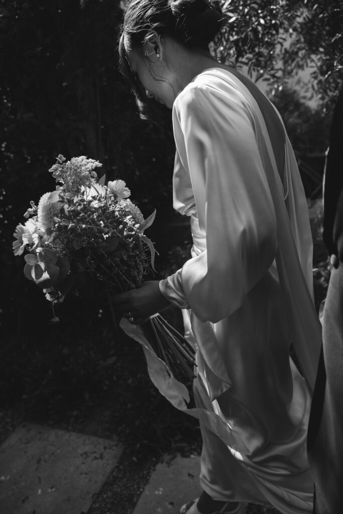 A couple in formal attire smiles and dances outdoors surrounded by greenery.