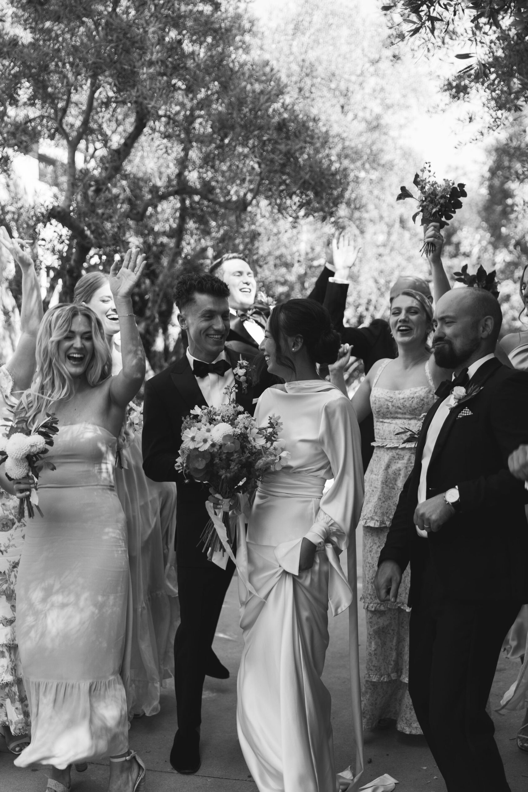 A wedding party celebrates outdoors, featuring the bride, groom, and several guests all in formal attire.