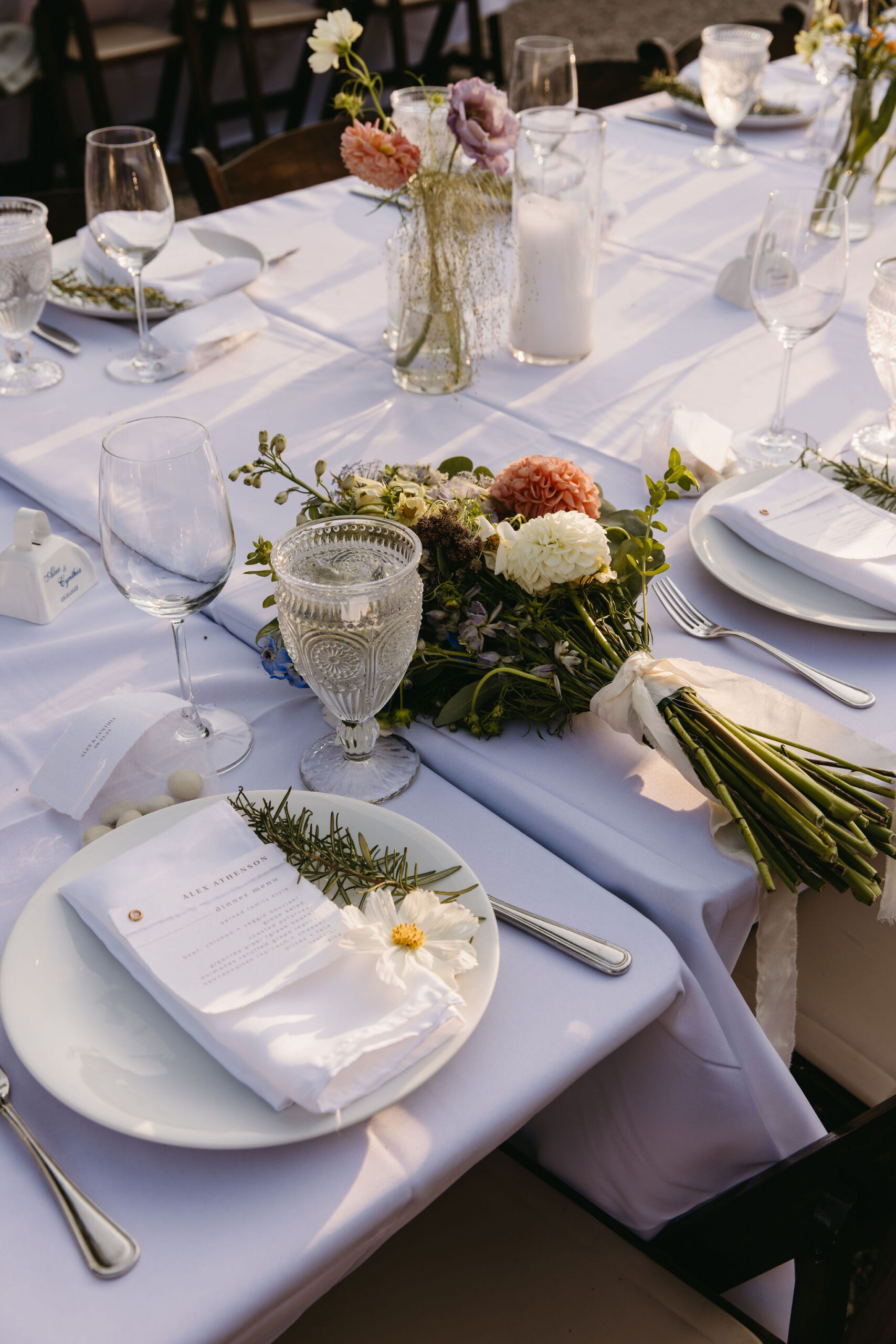 Table setting with white tablecloth, plates, glasses, and cutlery. A floral bouquet lies on the table alongside a menu card with a small flower on the plate. Candles and vases in the background.