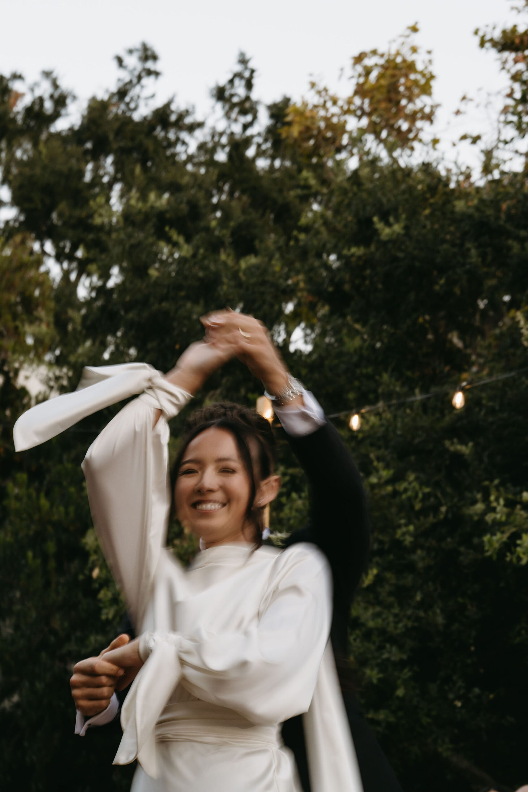 A bride and groom dance outdoors, smiling with trees and string lights in the background.