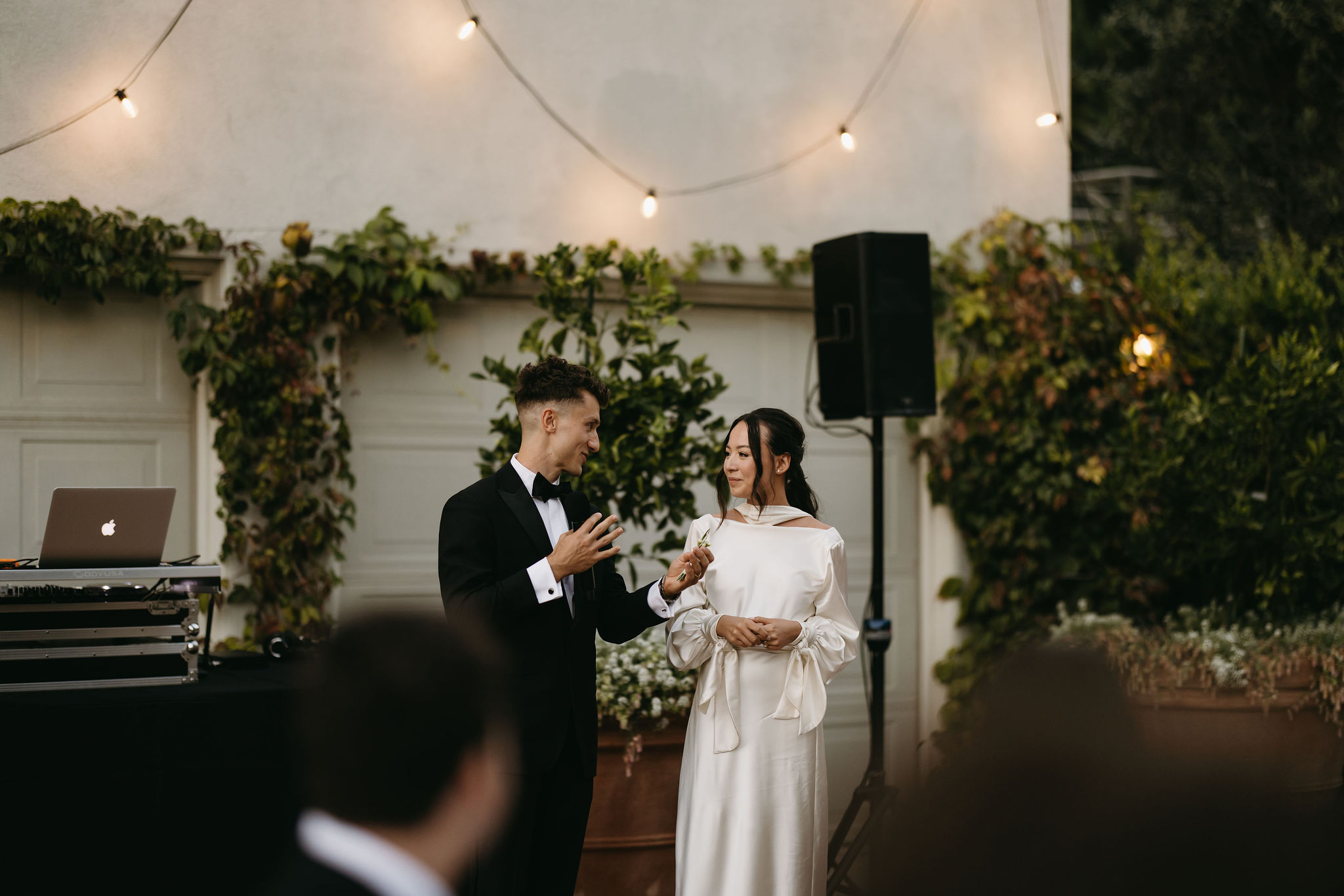 A couple in formal attire holds hands and stands near a DJ booth during an outdoor event, with string lights and greenery in the background.