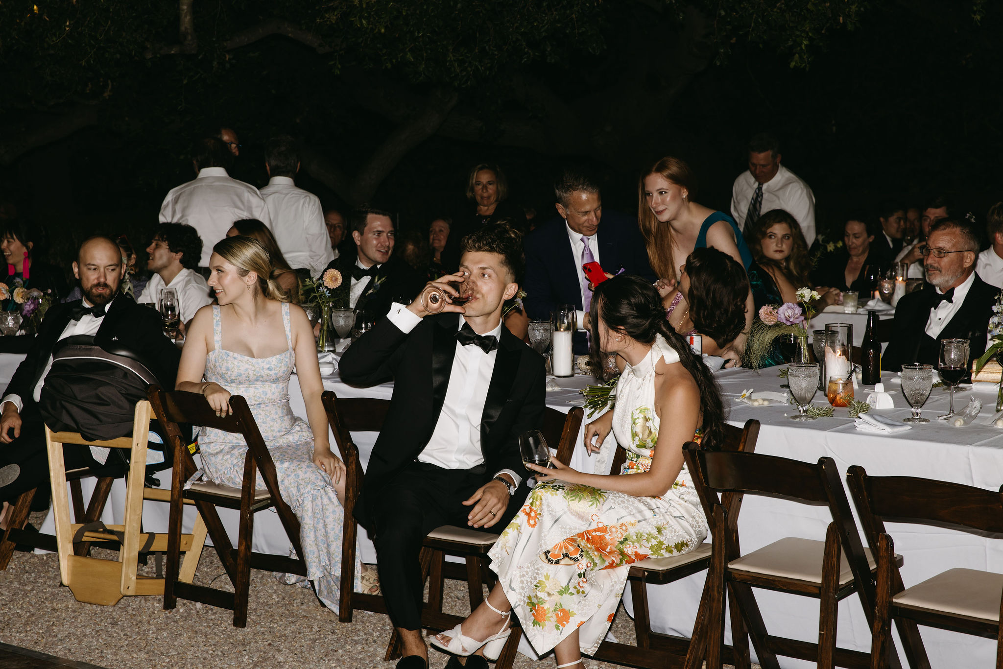 People seated at a long table outdoors, dressed in formal wear, engaging in conversation and dining.