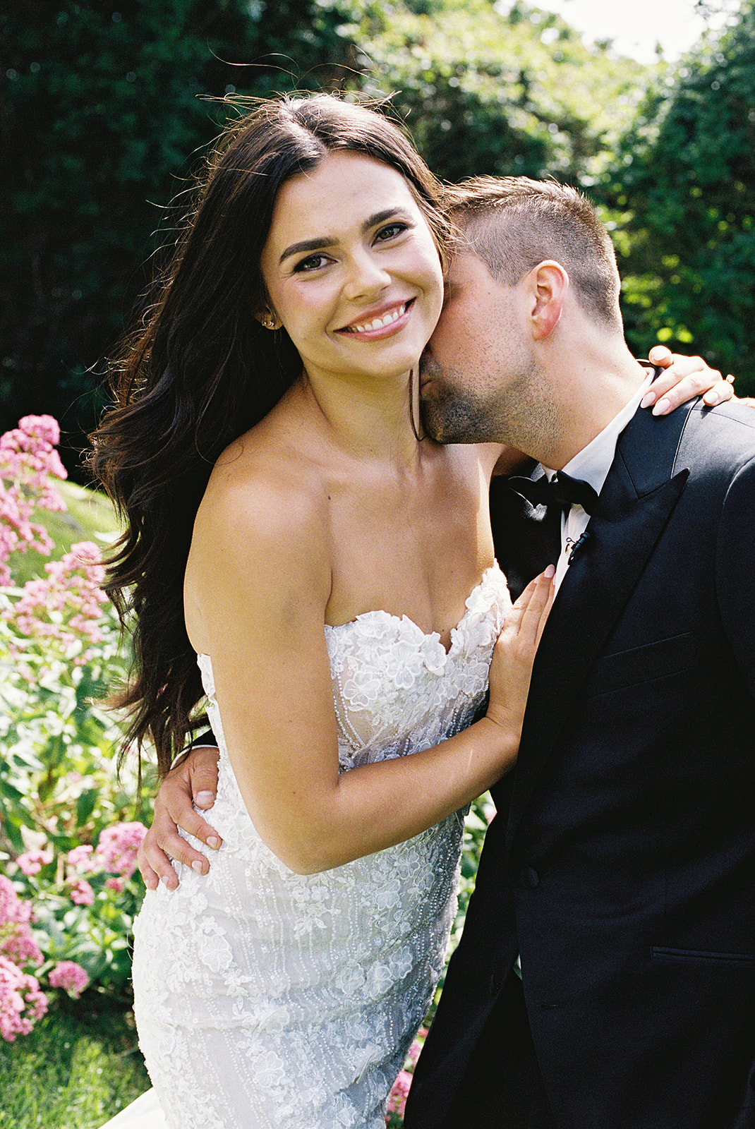 A woman in a white strapless dress smiles at the camera while a man in a suit kisses her cheek. They are outdoors with greenery and pink flowers in the background.