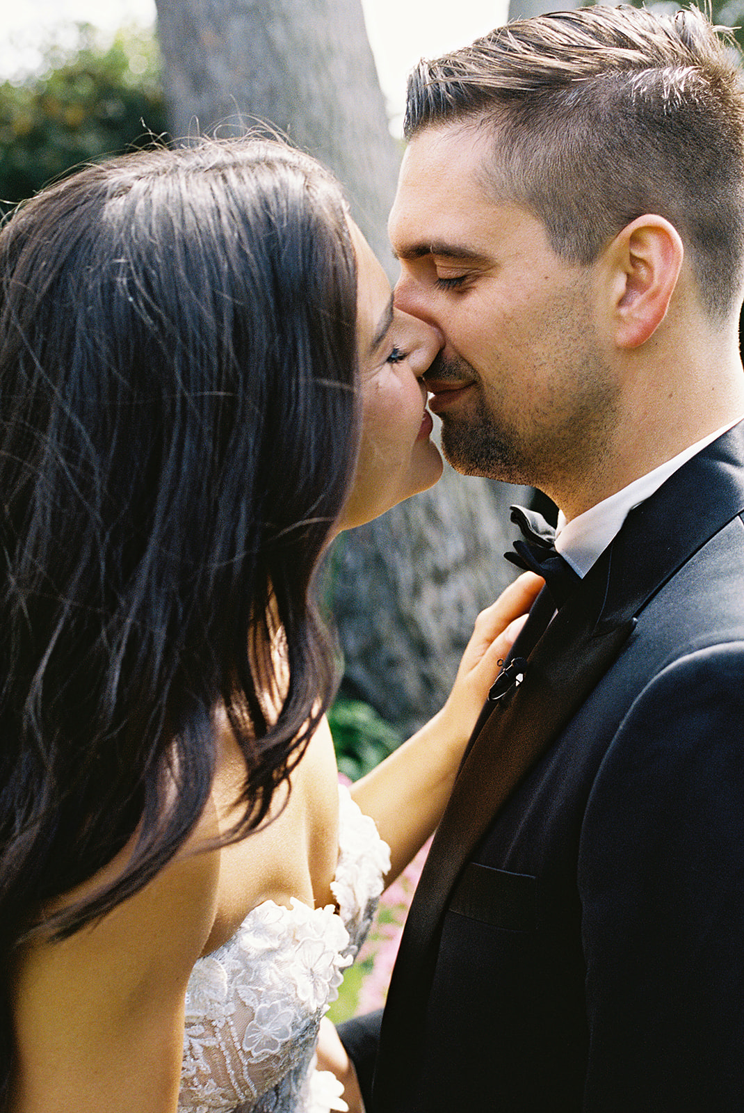 A couple kisses outdoors, dressed in formal wedding attire.