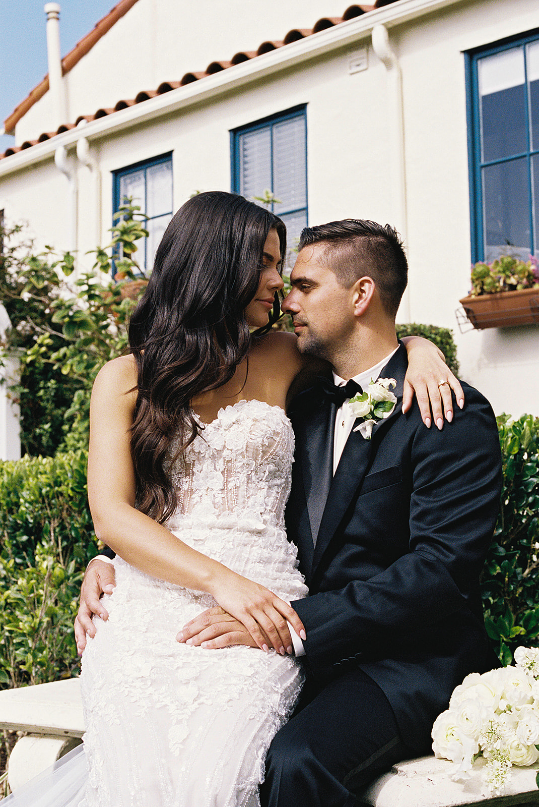 A bride in a white dress and a groom in a black suit stand facing each other in a garden, in front of a white house with blue-trimmed windows.