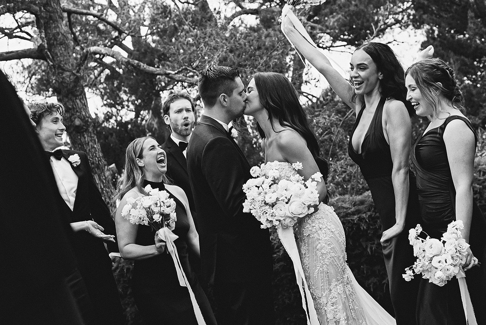 A bride and groom kiss in front of cheering bridesmaids and groomsmen, all dressed formally, in an outdoor setting.