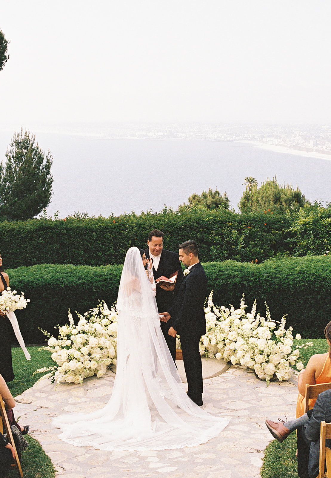 A couple stands before an officiant during an outdoor wedding ceremony with white flowers and a scenic background.