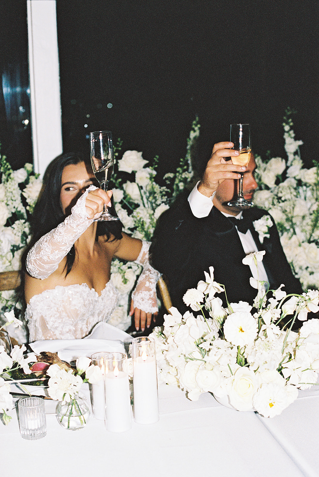 A couple sits at a table holding up wine glasses. The table is decorated with white flowers and candles.