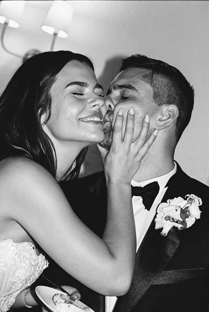 A bride in a wedding dress smiles while cupping the face of a groom in a tuxedo. They are both standing close together in a happy moment.