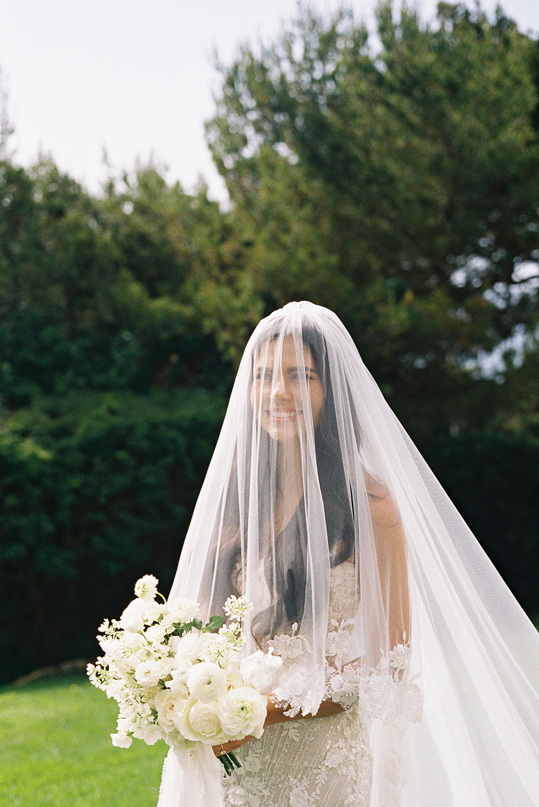 Bride wearing a white dress and veil holds a bouquet of white flowers, standing outdoors with greenery in the background.