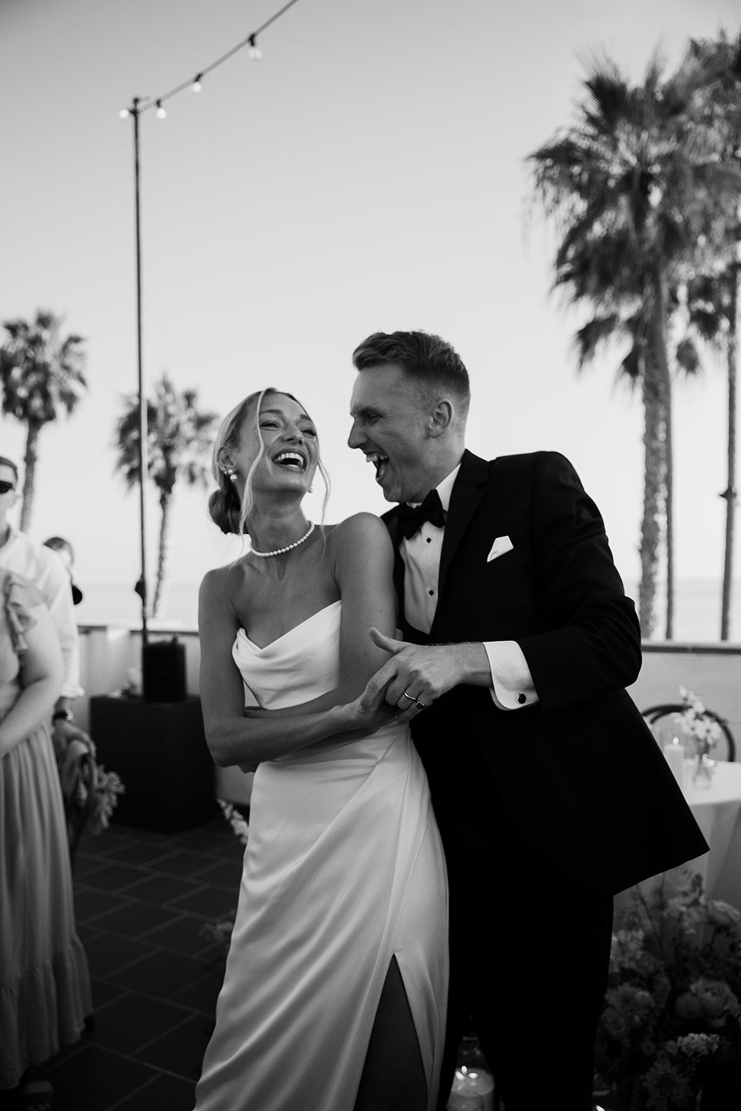 A couple dances closely at an outdoor event with tables and palm trees in the background. Guests watch them.