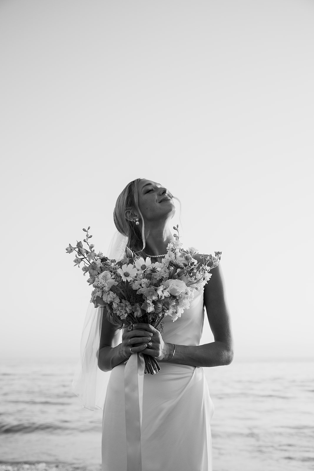 A bride in a white dress and veil holds a colorful bouquet, standing on a grassy area with tall palm trees in the background.