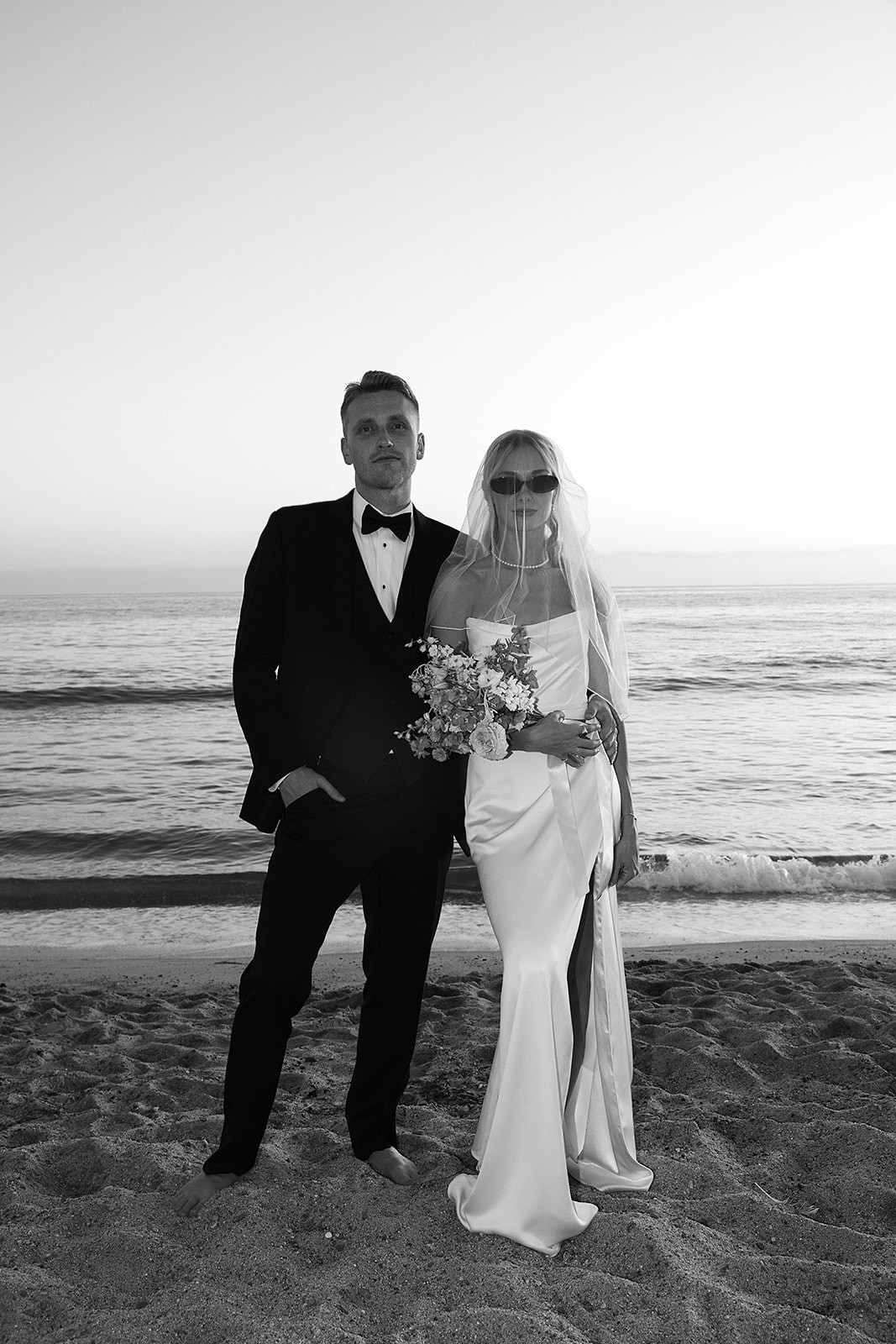 A bride in a white dress and veil holds a colorful bouquet, standing on a grassy area with tall palm trees in the background.