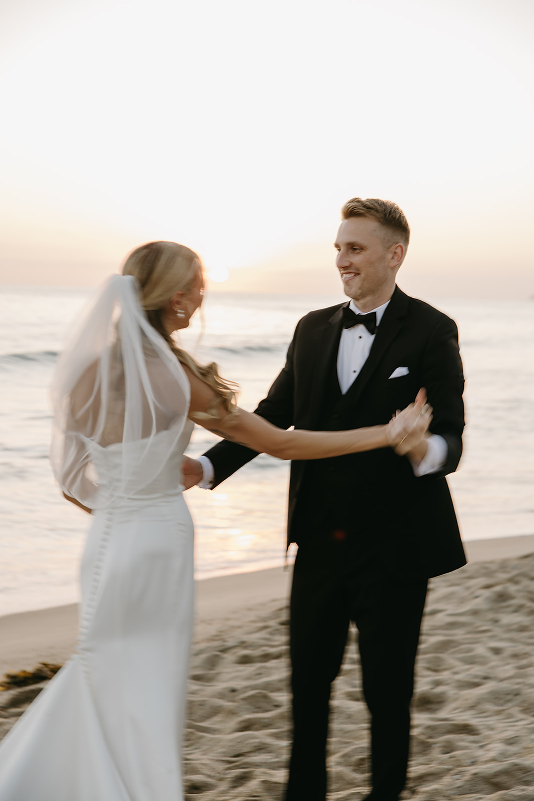 A bride in a white dress and veil holds a colorful bouquet, standing on a grassy area with tall palm trees in the background.