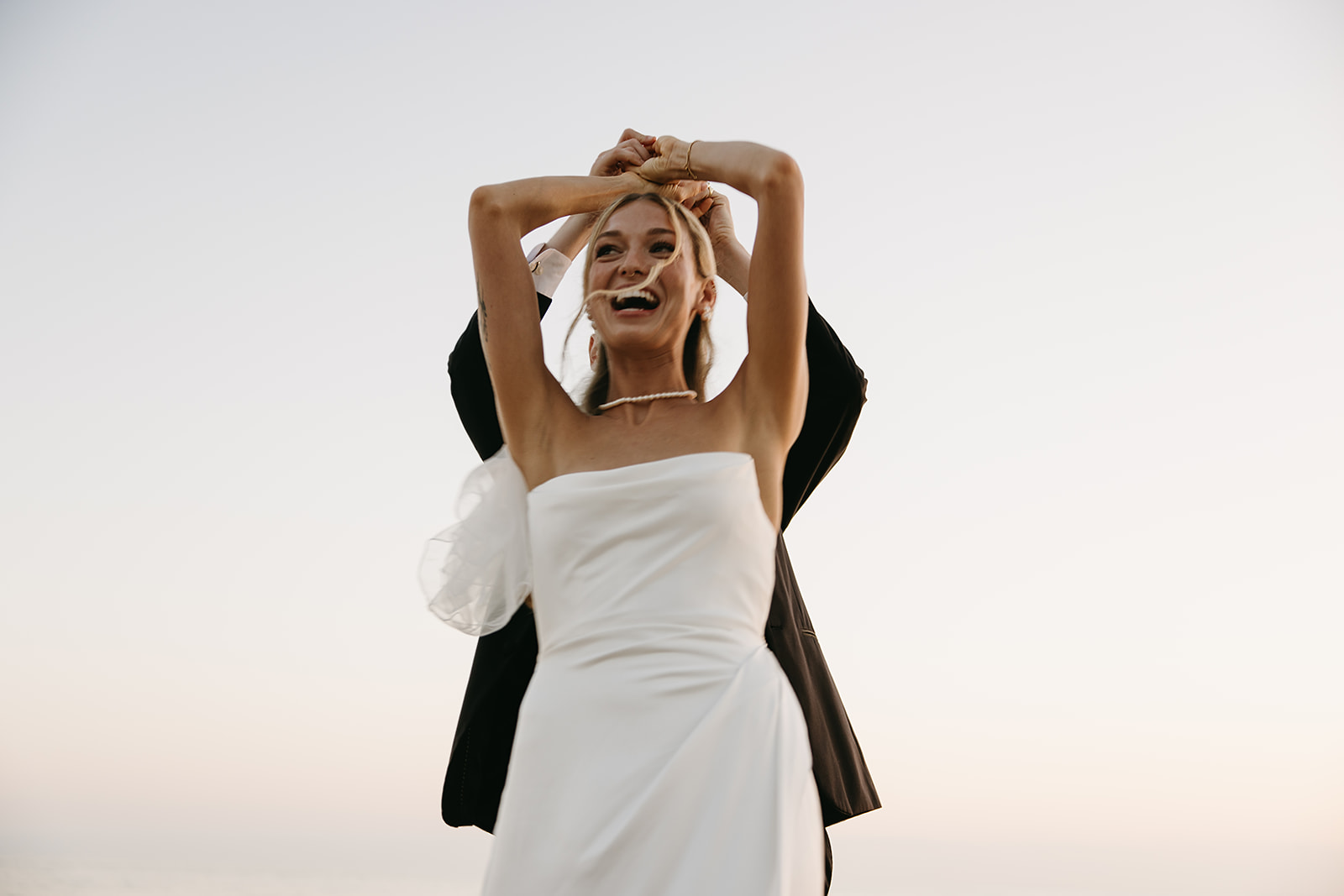 A bride in a white dress and veil holds a colorful bouquet, standing on a grassy area with tall palm trees in the background.