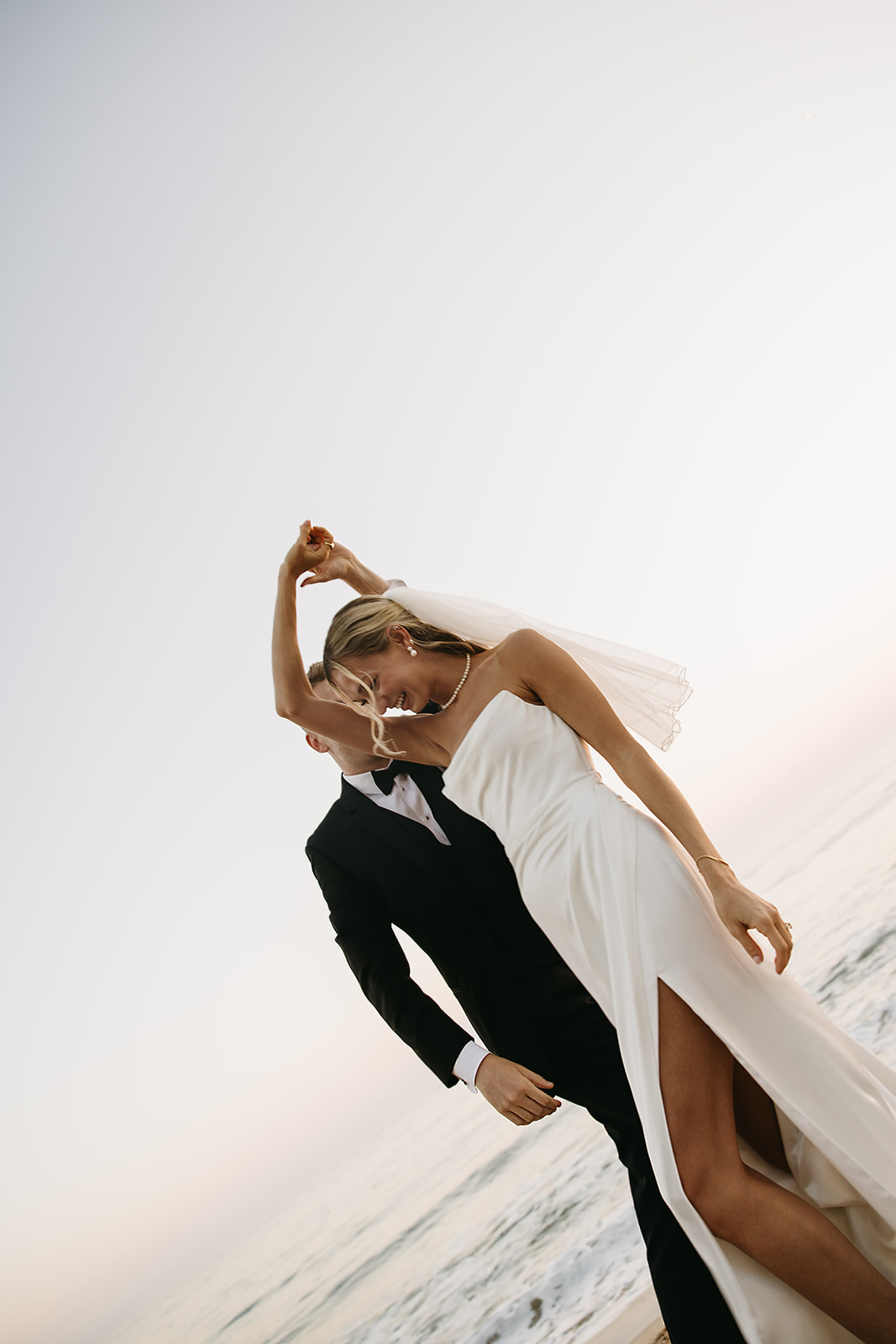 A bride in a white dress and veil holds a colorful bouquet, standing on a grassy area with tall palm trees in the background.