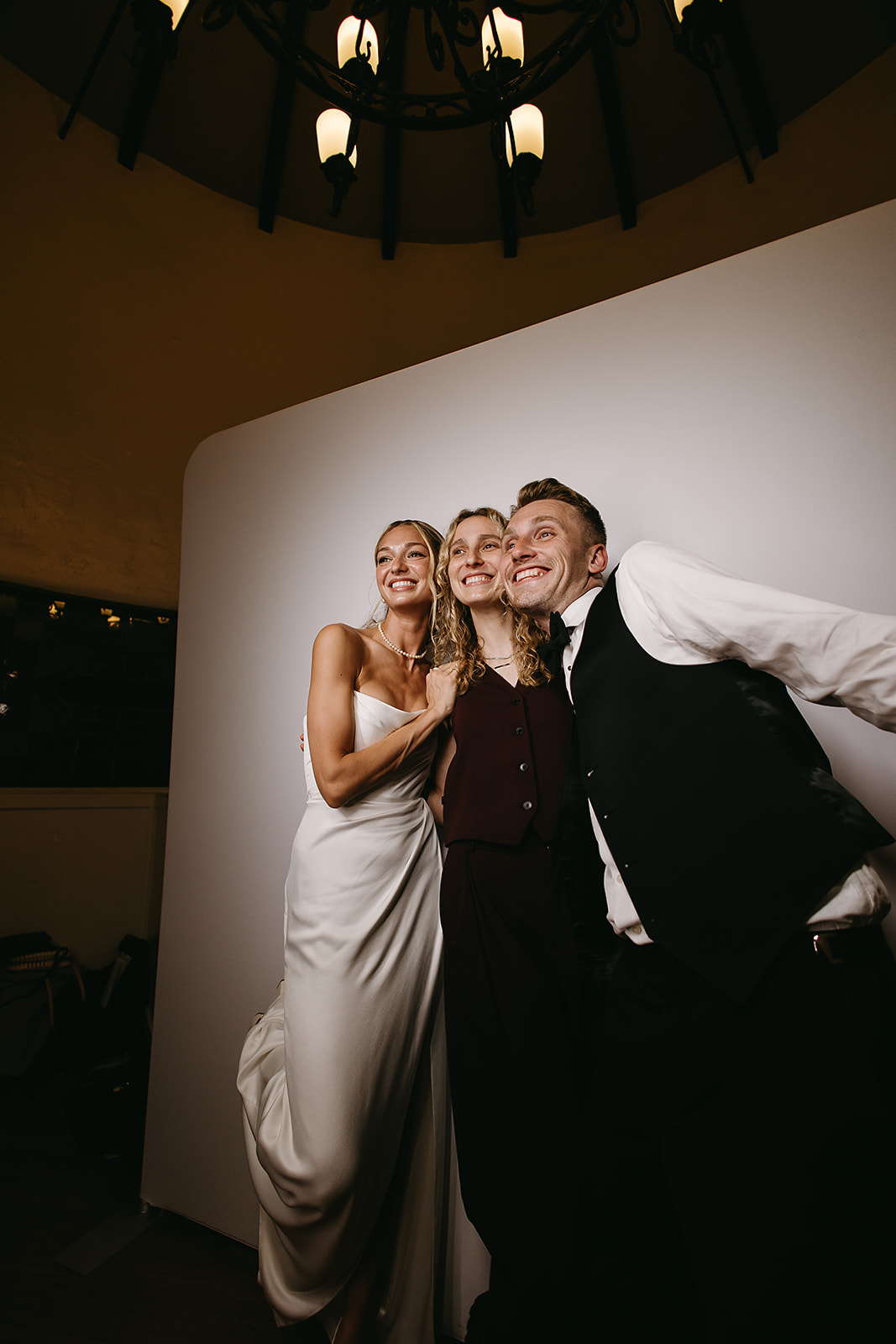 Three people, including one in a wedding dress and another in a suit, pose happily in front of a white backdrop under a chandelier.