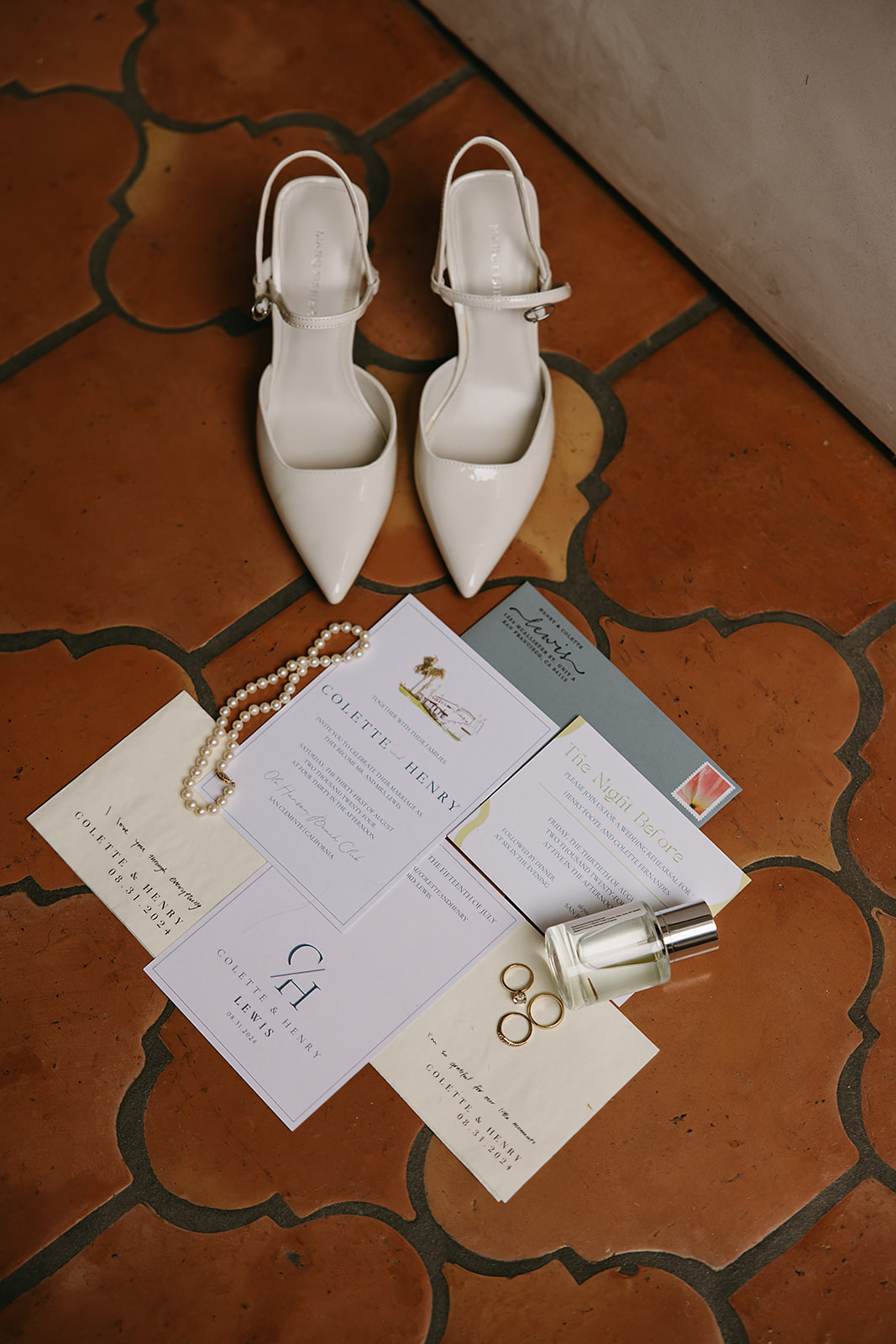 Wedding invitations, a pair of white shoes, pearl jewelry, three rings, a small perfume bottle, and floral cards arranged on a tiled floor.