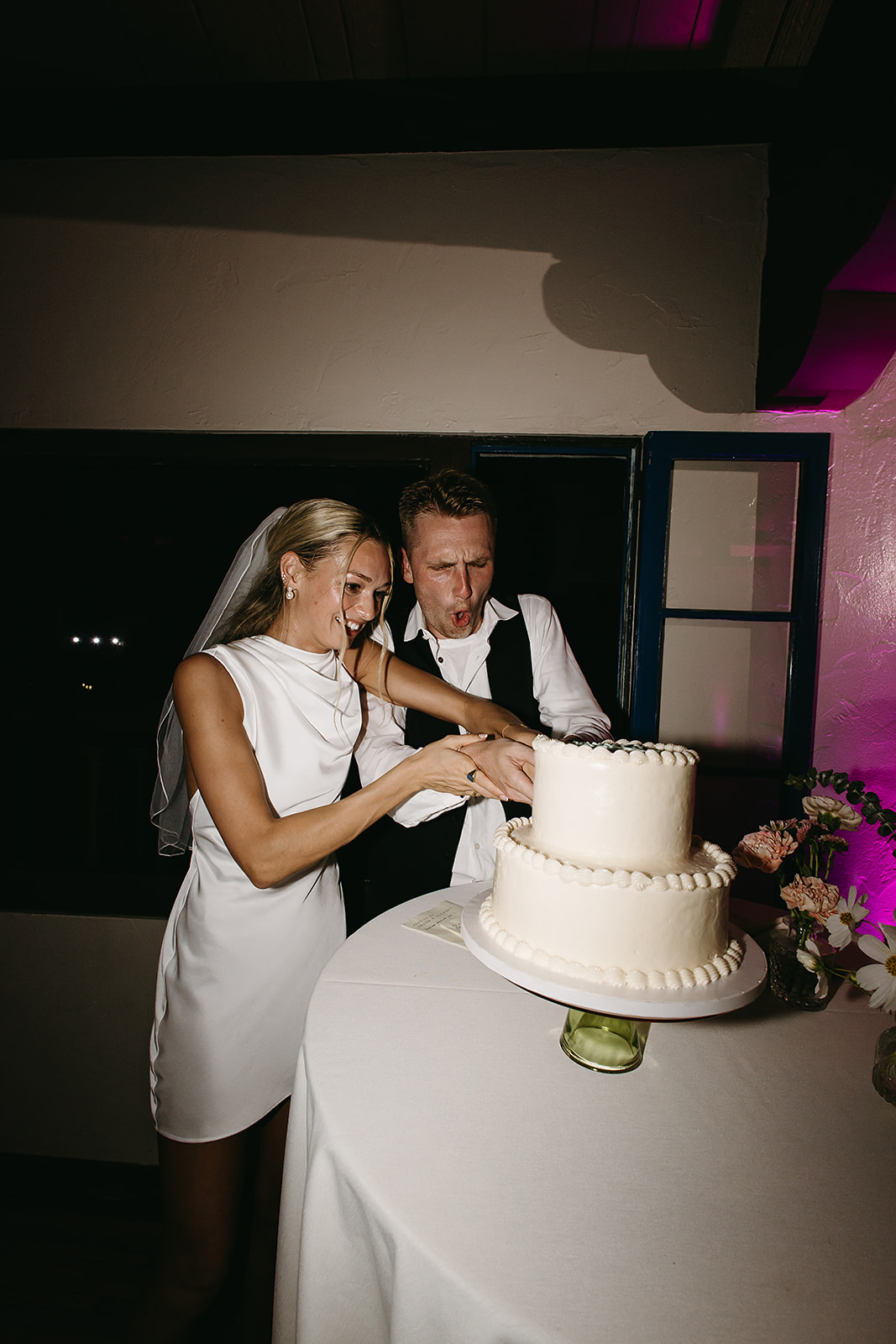 A bride and groom are cutting a white tiered cake at their wedding reception. Both are smiling and standing next to a table with a floral arrangement.