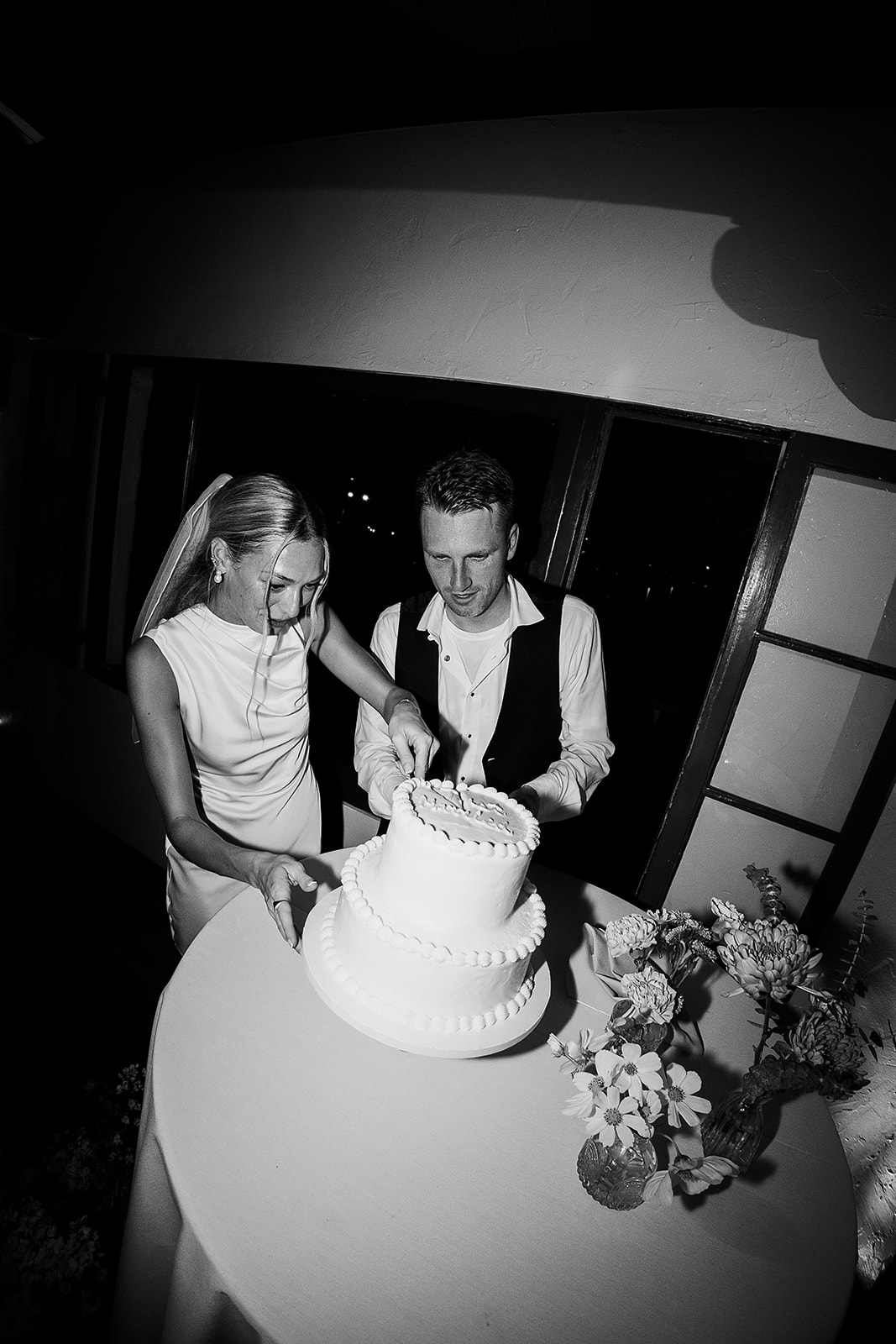 A bride and groom are cutting a white tiered cake at their wedding reception. Both are smiling and standing next to a table with a floral arrangement.